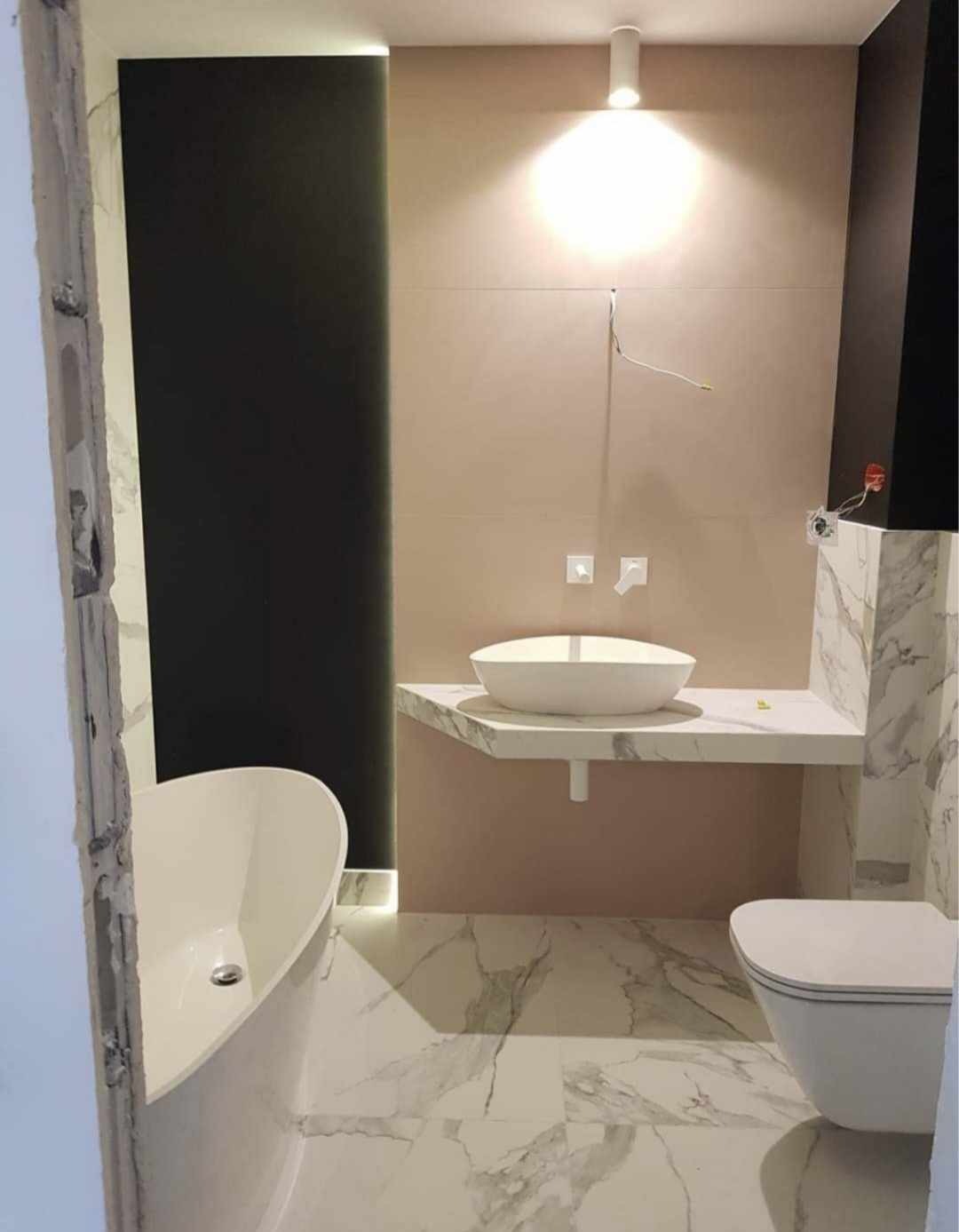 Nowoczesna łazienka z marmurową podłogą, wolnostojącą wanną i umywalką nablatową na blacie z tego samego materiału, oświetlona punktowym światłem, z widocznymi niedokończonymi elementami...