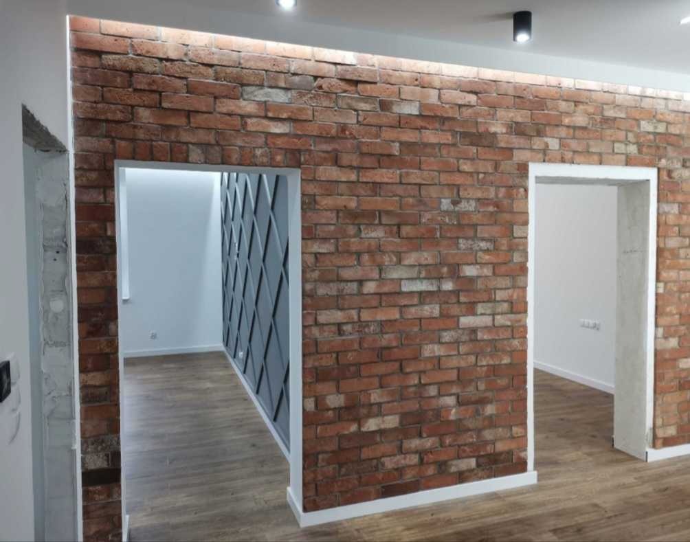 Ściana z czerwonej cegły z dwoma otworami drzwiowymi, jeden z widoczną ścianą z panelami dekoracyjnymi w romby, drewniana podłoga, oświetlenie sufitowe.