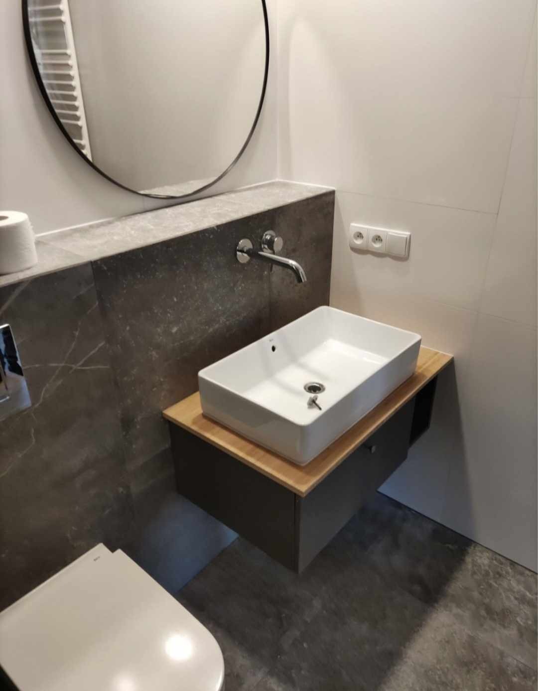 Nowoczesna łazienka z prostokątną umywalką nablatową, szarą szafką pod umywalką z drewnianym blatem, okrągłym lustrem i marmurową ścianą.