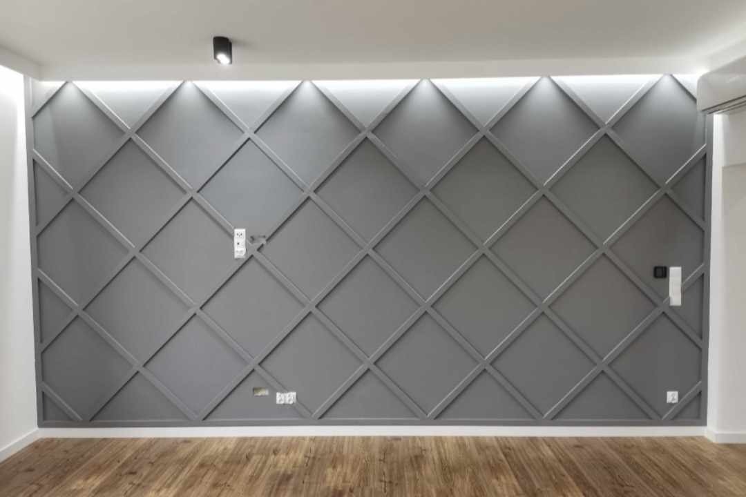 Szara ściana z dekoracyjnymi listwami tworzącymi romboidalny wzór, oświetlona od góry taśmą LED, widoczne gniazdka elektryczne i drewnopodobna podłoga.