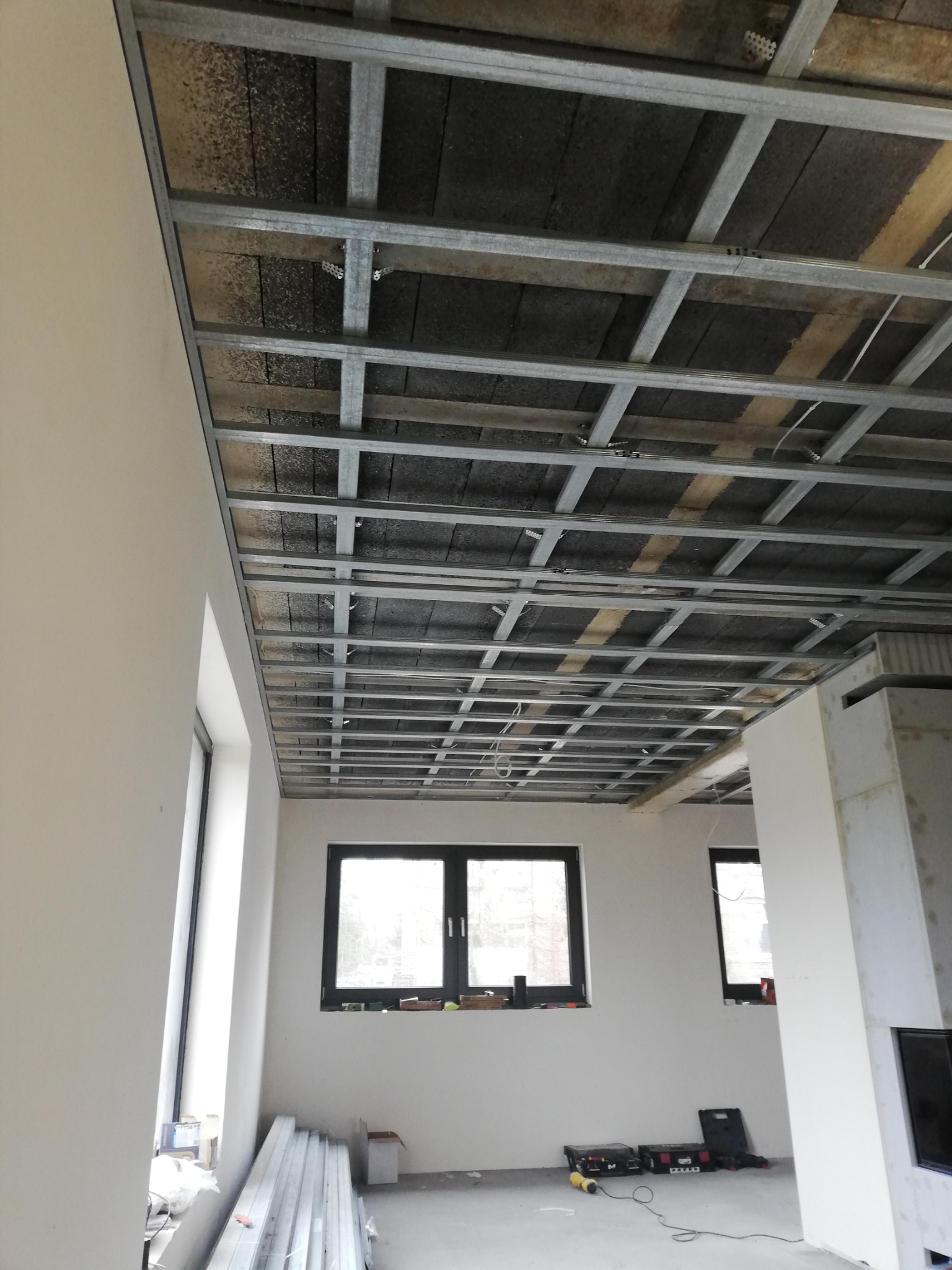 Metalowa konstrukcja sufitu podwieszanego z izolacją, widok z dołu w trakcie montażu, narzędzia i profile na podłodze, dwa okna z czarnymi ramami w tle.