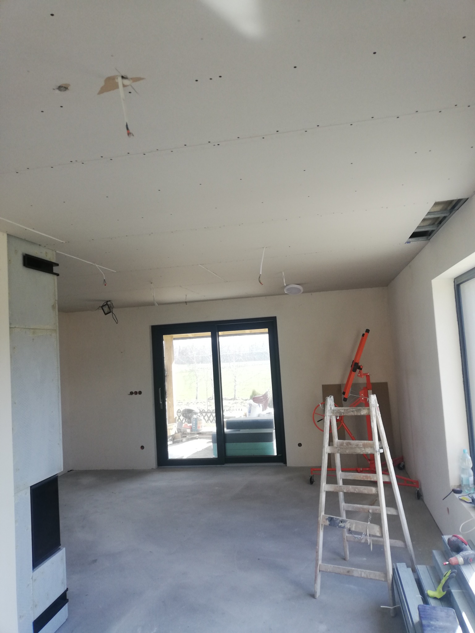 Wnętrze w trakcie wykańczania: sufit z płyt kartonowo-gipsowych z widocznymi punktami mocowań, drabina oparta o ścianę, podnośnik do płyt, częściowo zabudowany kominek i duże okno przesuwne.