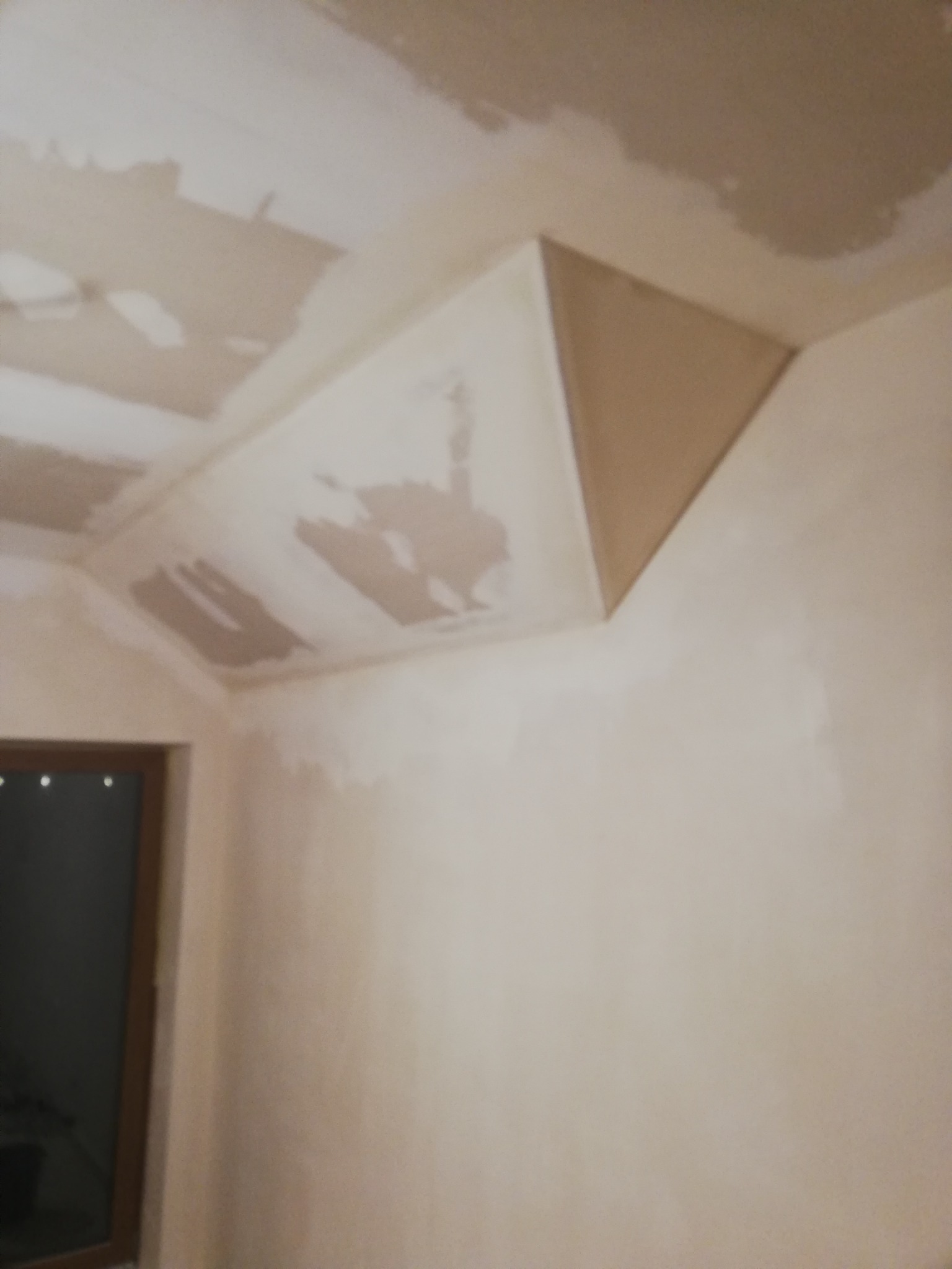 Niedokończone wykończenie sufitu i ściany w pomieszczeniu z widocznym oknem, ślady gładzi szpachlowej i narożnik z płyty gipsowej.