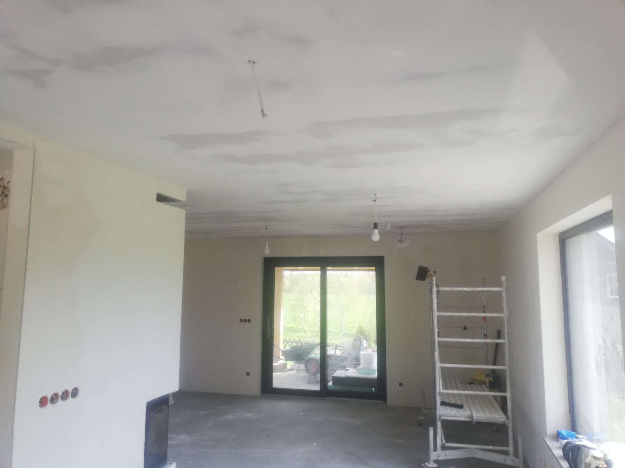 Wnętrze w trakcie prac wykończeniowych: niedokończone gładzie na suficie, widoczna drabina malarska, surowe ściany z puszkami elektrycznymi, duże okno przesuwne z widokiem na zewnątrz.