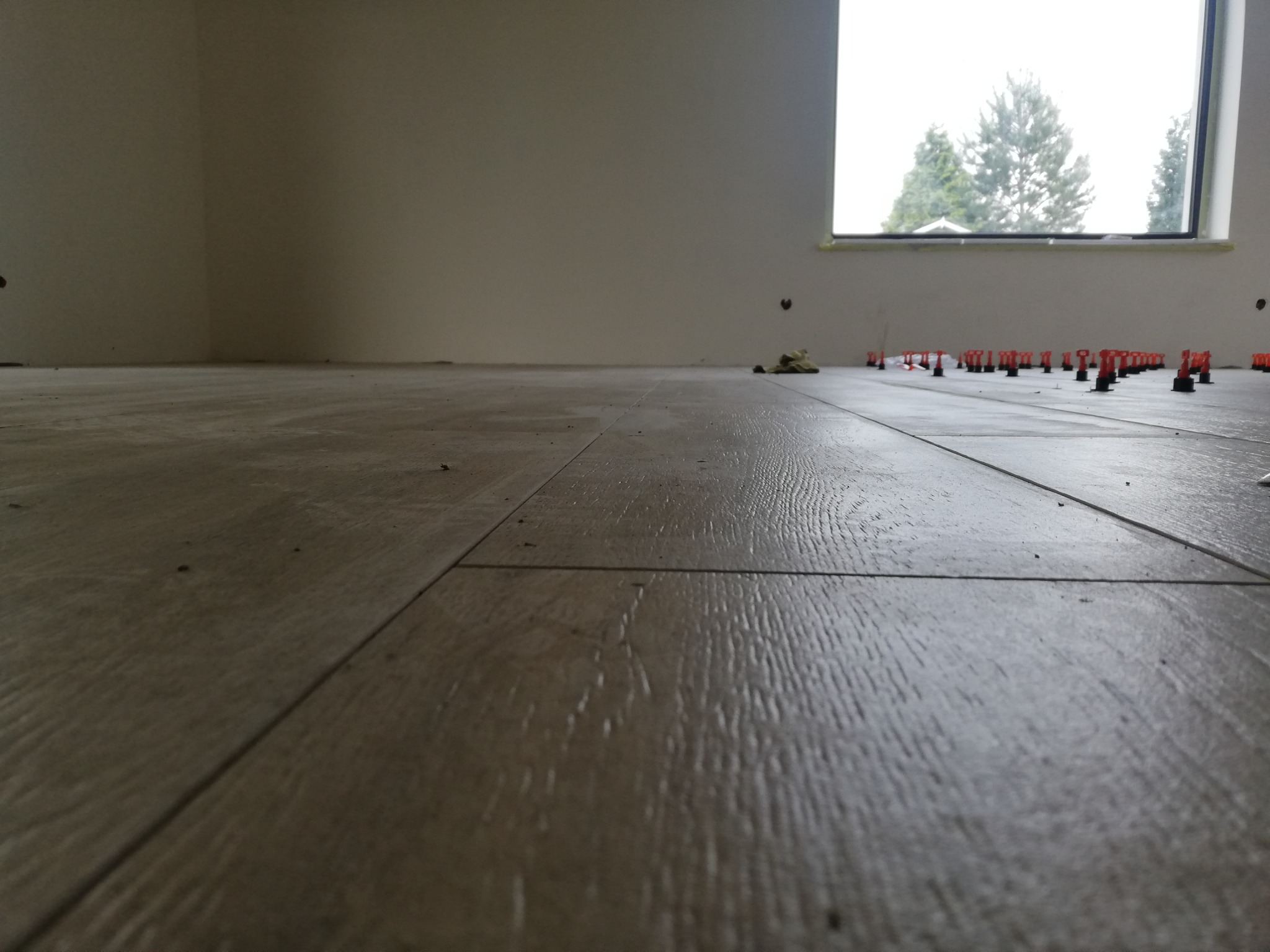 Układanie paneli podłogowych z widocznymi elementami poziomującymi, w tle okno z widokiem na drzewa.