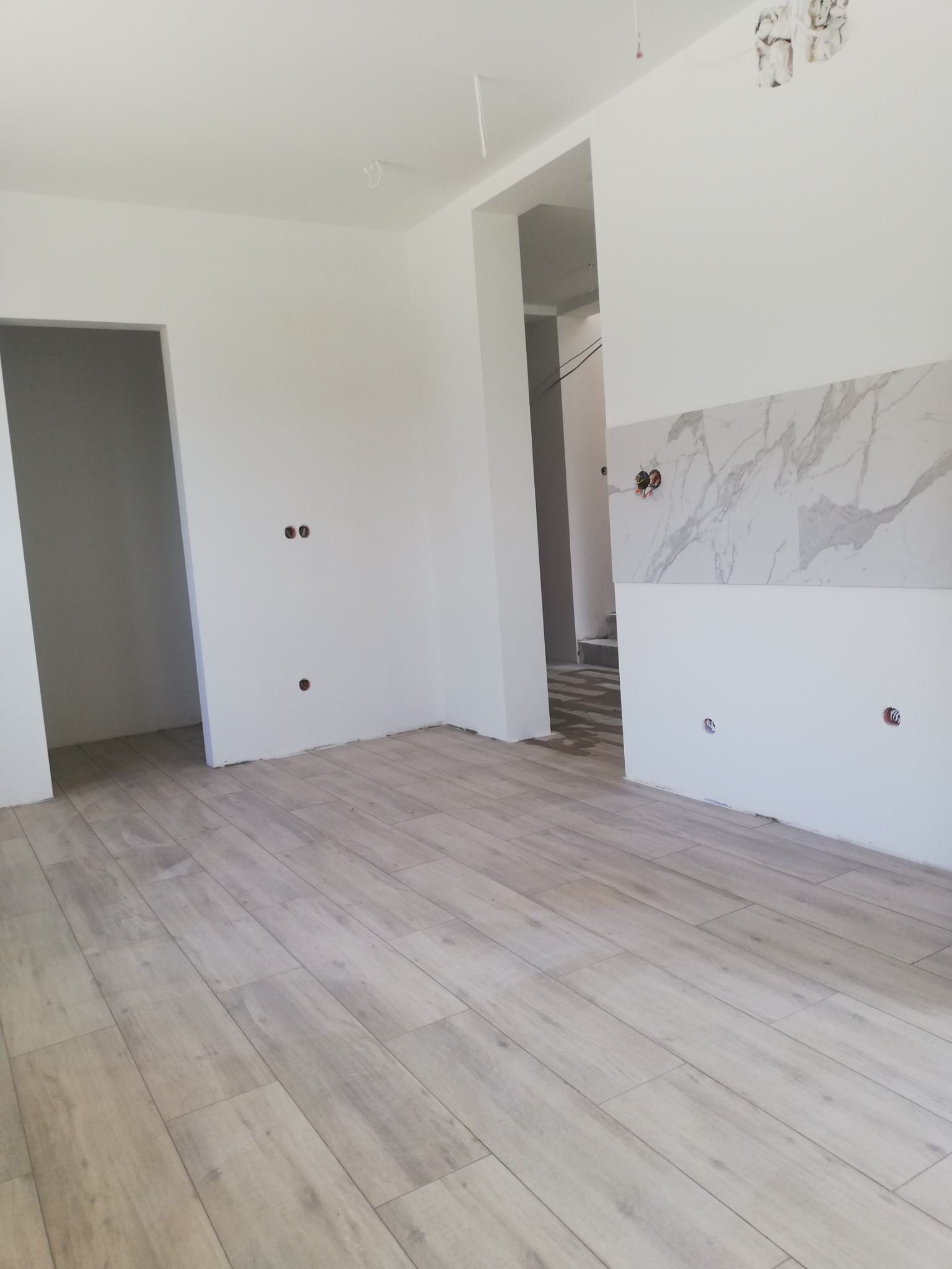 Jasne panele podłogowe w nowym mieszkaniu, fragment ściany z imitacją marmuru, przygotowane gniazdka elektryczne, widoczne ślady po kuciu w ścianie.