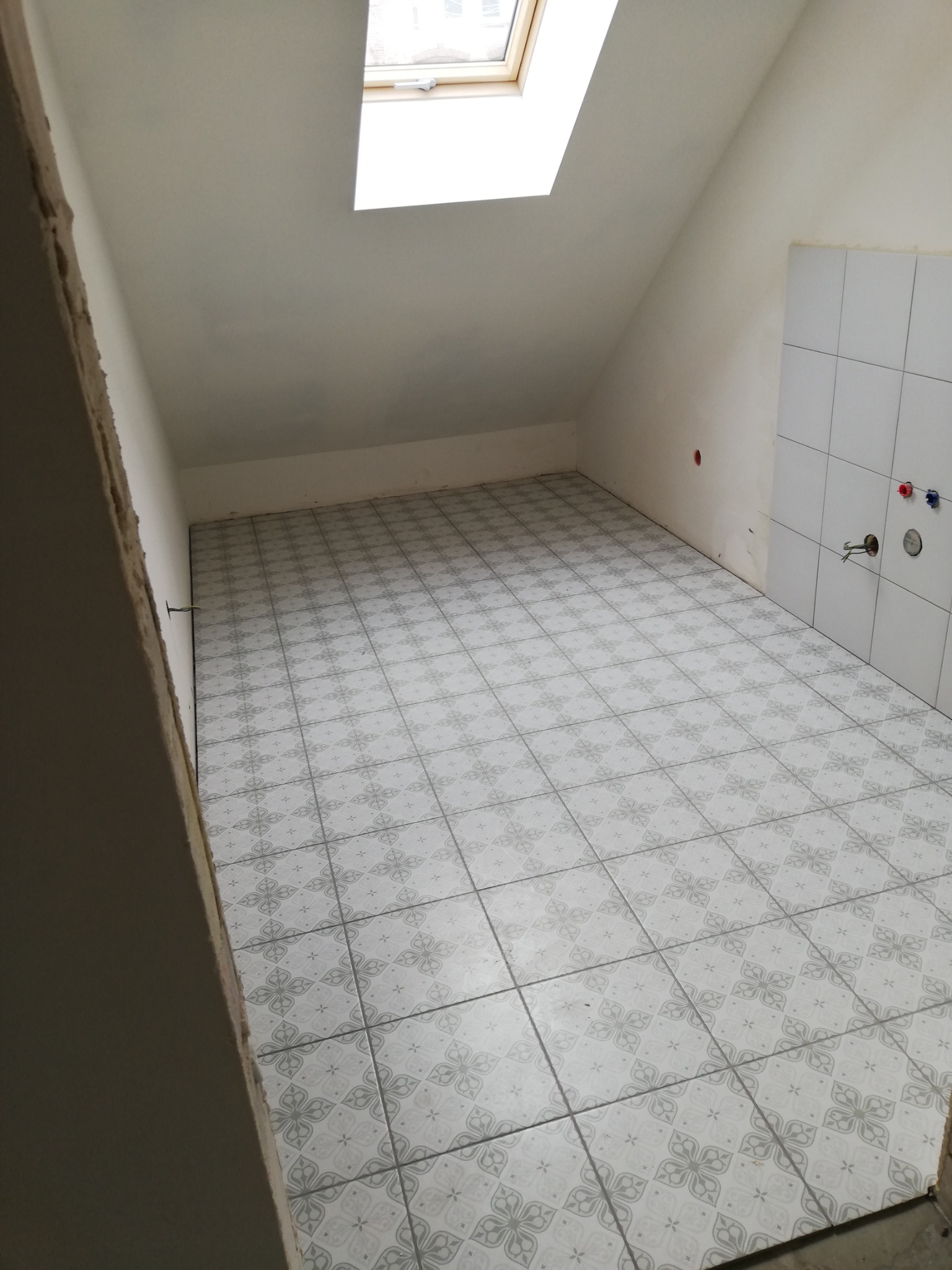 Poddasze po wyłożeniu podłogi jasnymi, wzorzystymi płytkami; widoczne fragmenty ścian z instalacją wodną i okno dachowe.