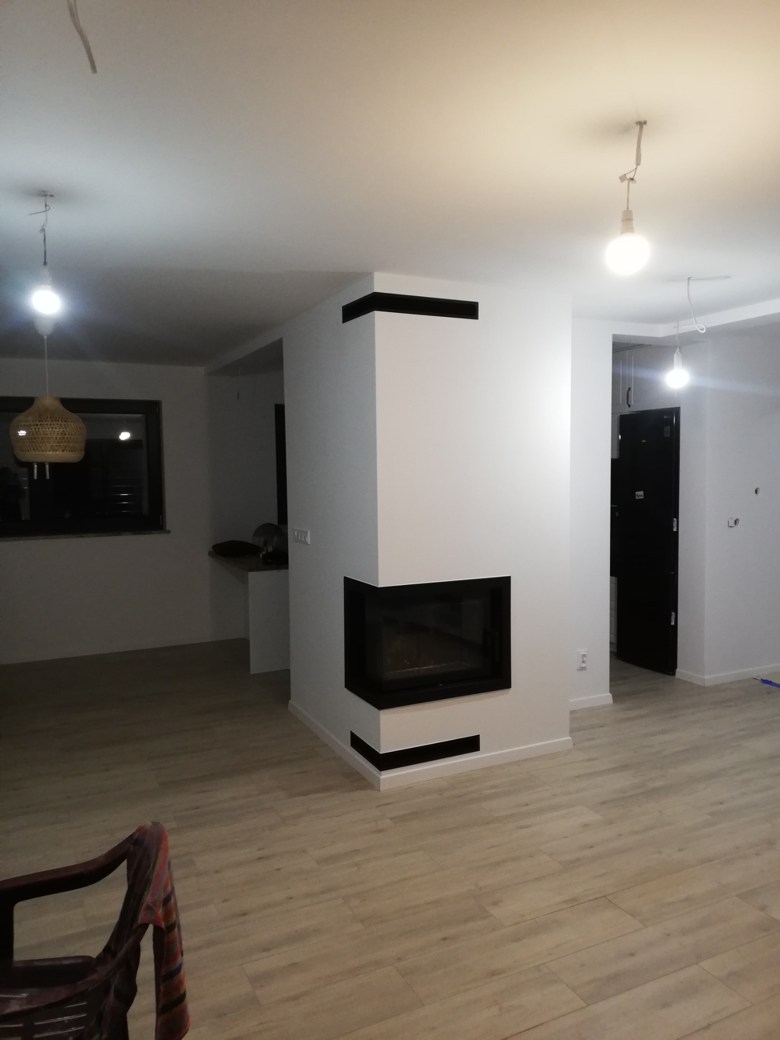 Salon po remoncie z nowoczesnym kominkiem obudowanym białą konstrukcją z czarnymi akcentami, drewnopodobna podłoga i wiszące lampy bez kloszy.