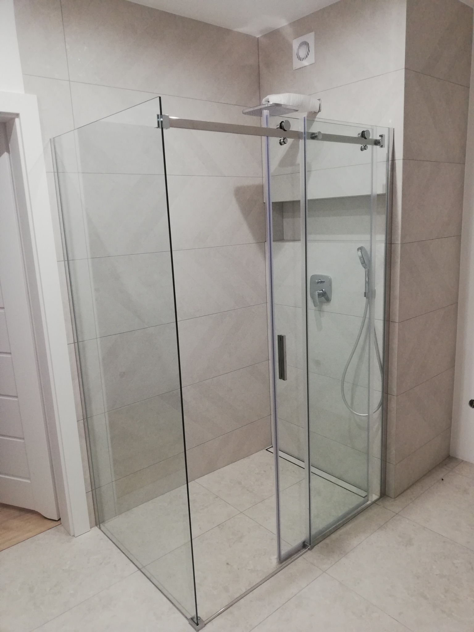 Nowoczesna kabina prysznicowa z przesuwnymi drzwiami, wykończona jasnymi płytkami w neutralnych odcieniach, z widoczną armaturą i odpływem liniowym w podłodze.