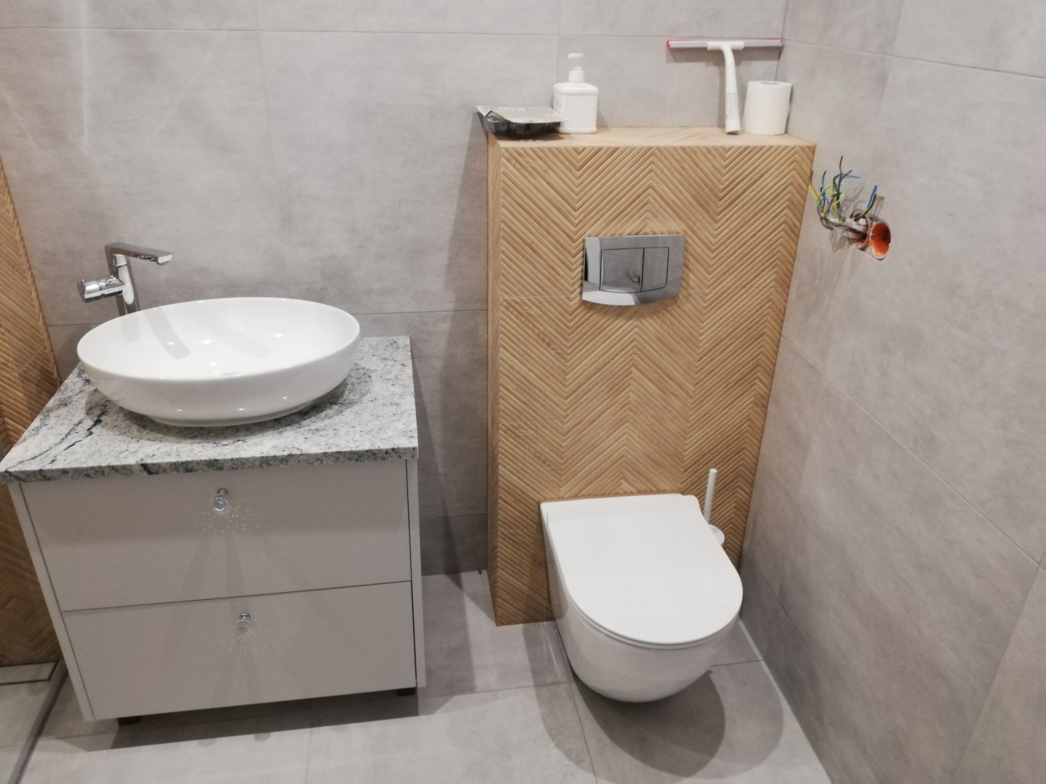 Nowoczesna łazienka z umywalką nablatową na szafce z blatem imitującym granit, wiszącą toaletą i drewnianą zabudową stelaża z wzorem jodełki, na ścianie widoczne wystające kable.