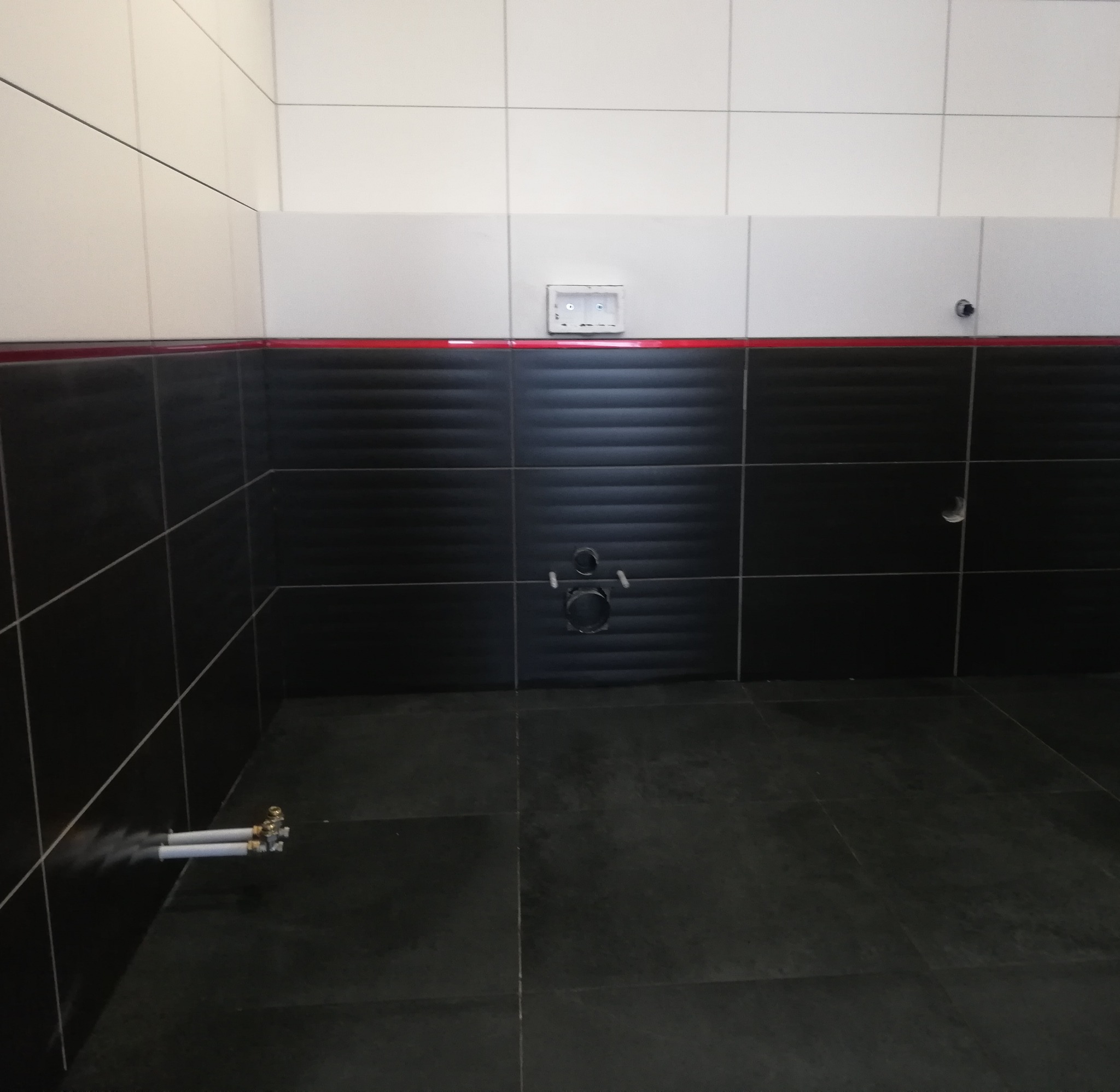 Czarne i białe płytki ścienne w łazience, z widocznymi rurami wodnymi i gniazdem elektrycznym w trakcie instalacji.