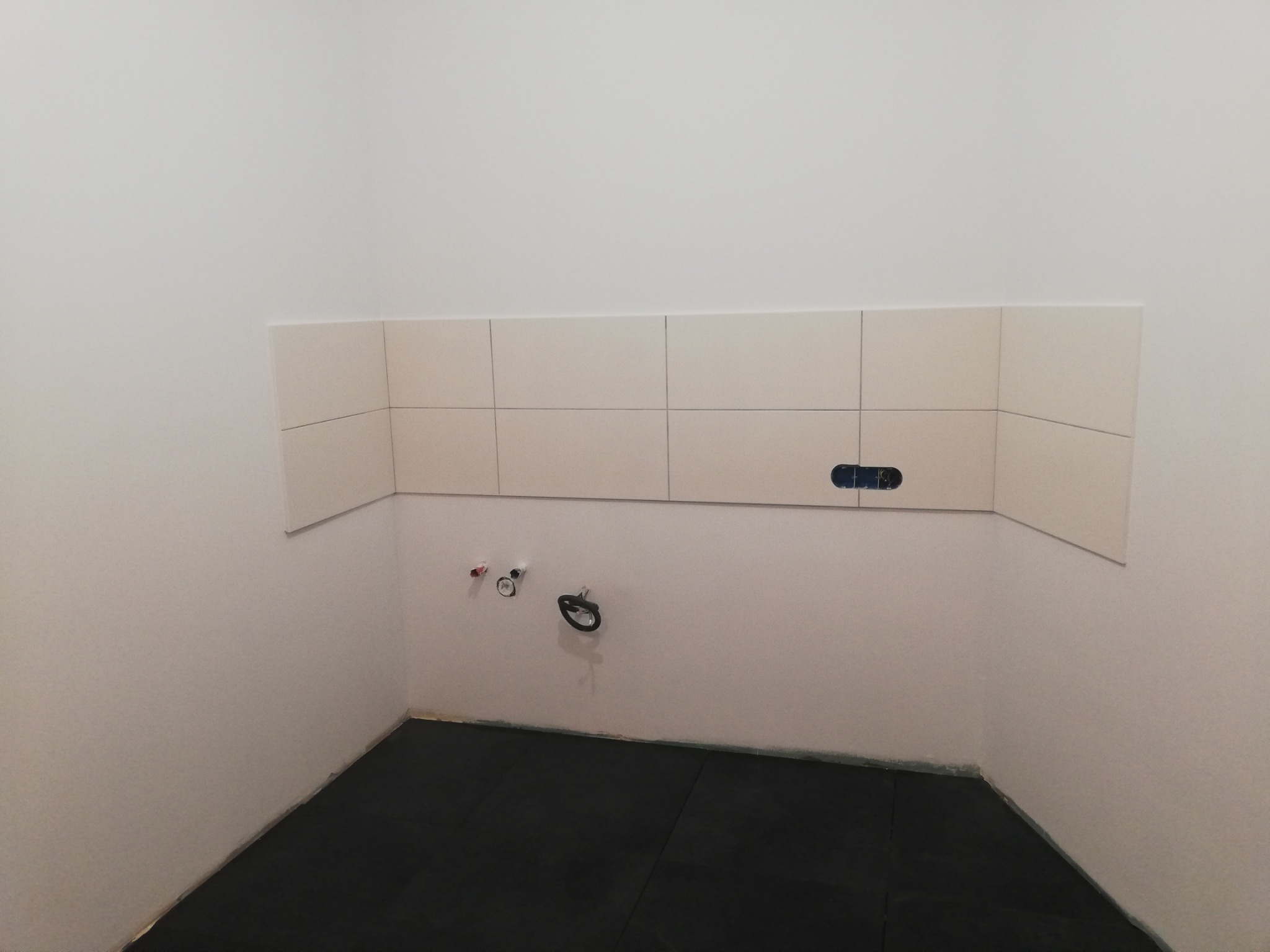 Fragment ściany w trakcie wykańczania z częściowo położonymi jasnymi płytkami ceramicznymi, widoczne przyłącza wodne i czarna podłoga z gumy.