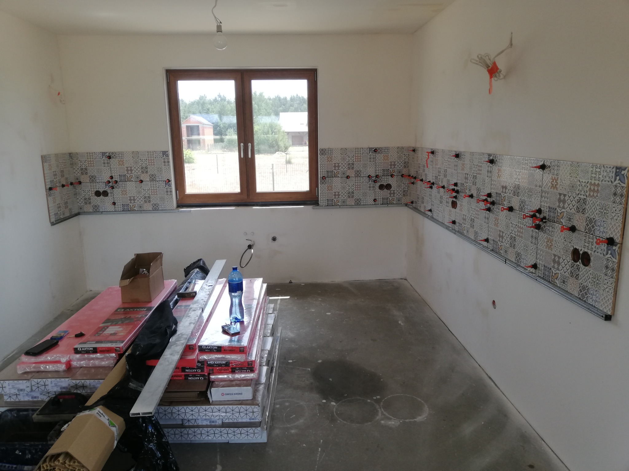 Fragment ściany z częściowo ułożonymi płytkami dekoracyjnymi w kuchni, z widocznymi krzyżykami dystansowymi i puszkami elektrycznymi, na podłodze stosy opakowań płytek i butelka wody.