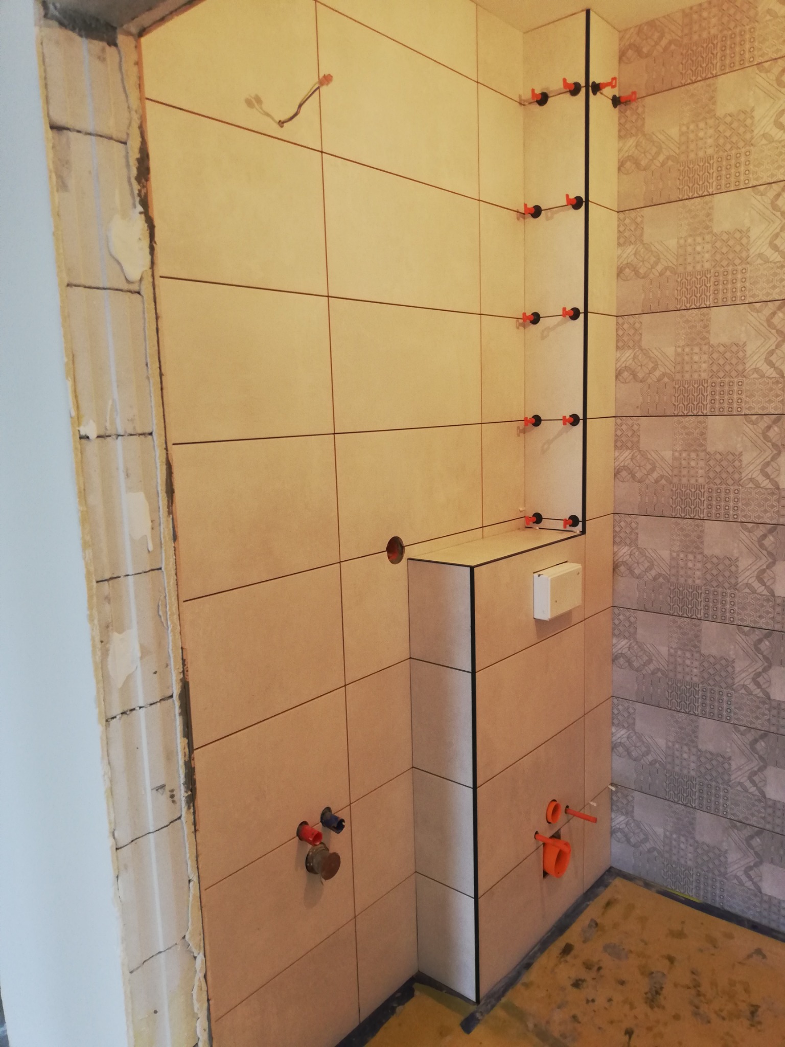 Ściany łazienki w trakcie wykańczania: beżowe płytki ścienne ułożone do pewnej wysokości, widoczne elementy instalacji wodnej i kanalizacyjnej, system poziomowania płytek w narożniku, surowy stan...