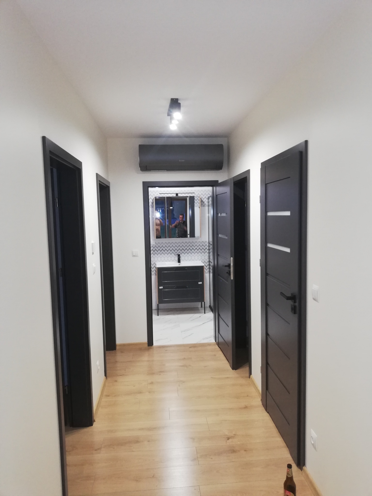 Korytarz po remoncie z trzema parami czarnych drzwi, jasnymi panelami podłogowymi i widoczną łazienką z szaro-białą mozaiką nad umywalką. Na ścianie zamontowany klimatyzator.