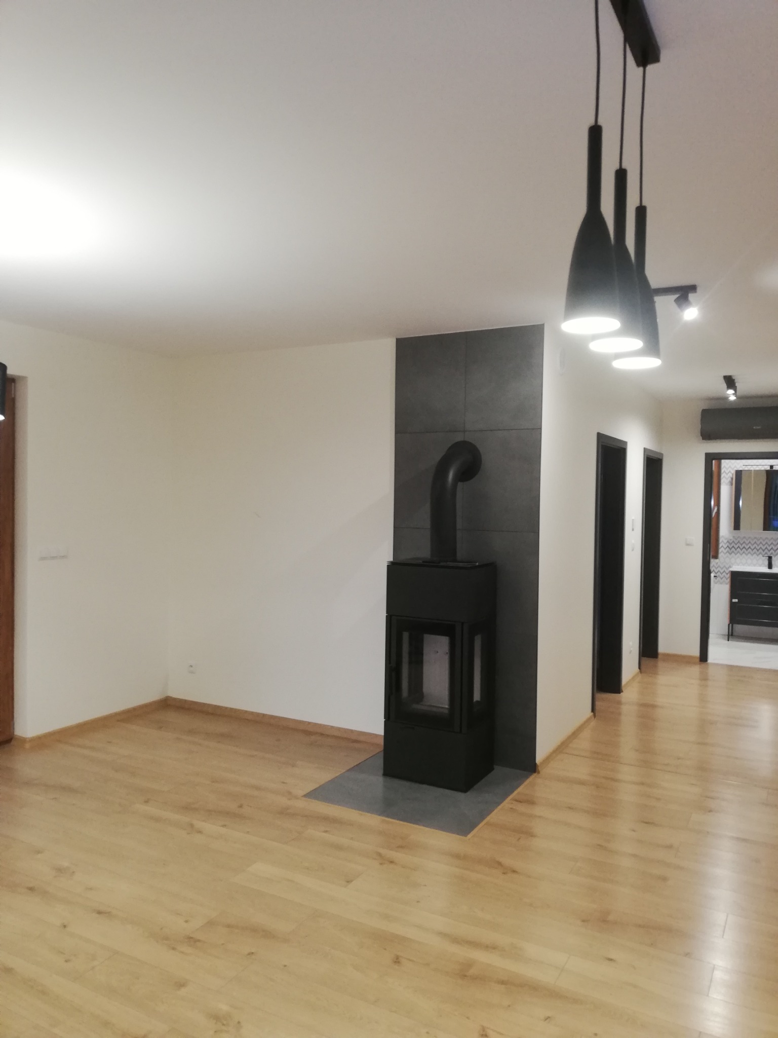 Wnętrze salonu z jasną podłogą panelową i nowoczesnym, czarnym piecem wolnostojącym obudowanym szarą płytą, wiszące lampy w stylu industrialnym.