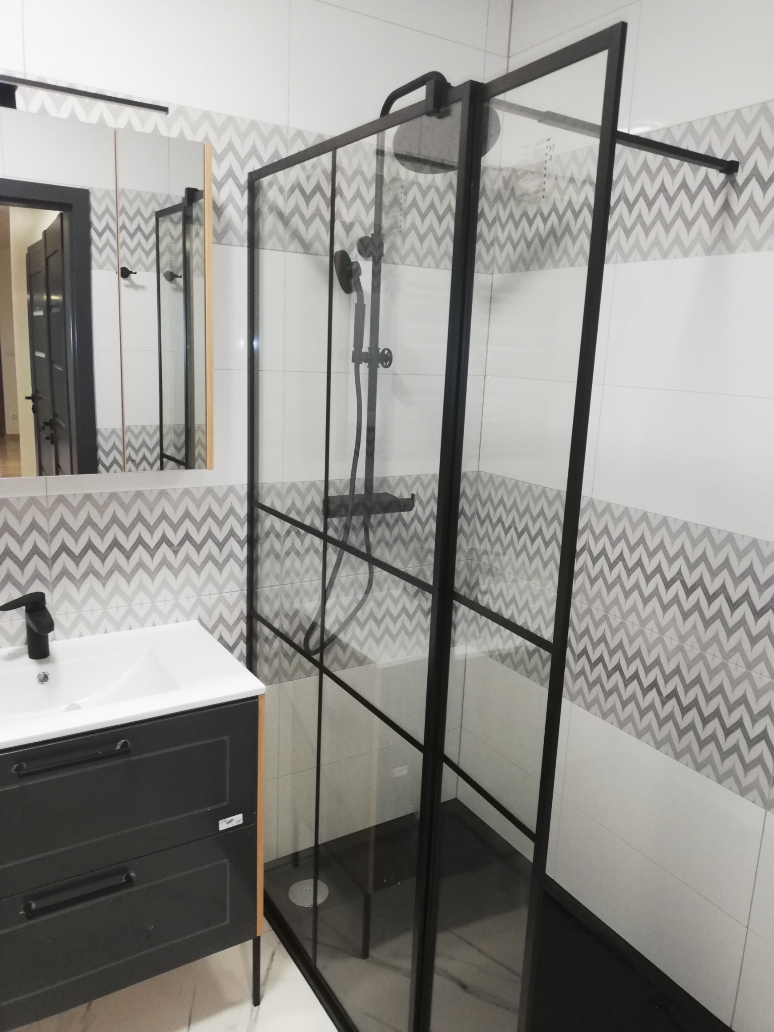 Nowoczesna kabina prysznicowa z czarnymi profilami i czarnym brodzikiem w łazience w Świebodzicach, z umywalką na szafce w kolorze antracytowym i wzorzystymi płytkami na ścianach.