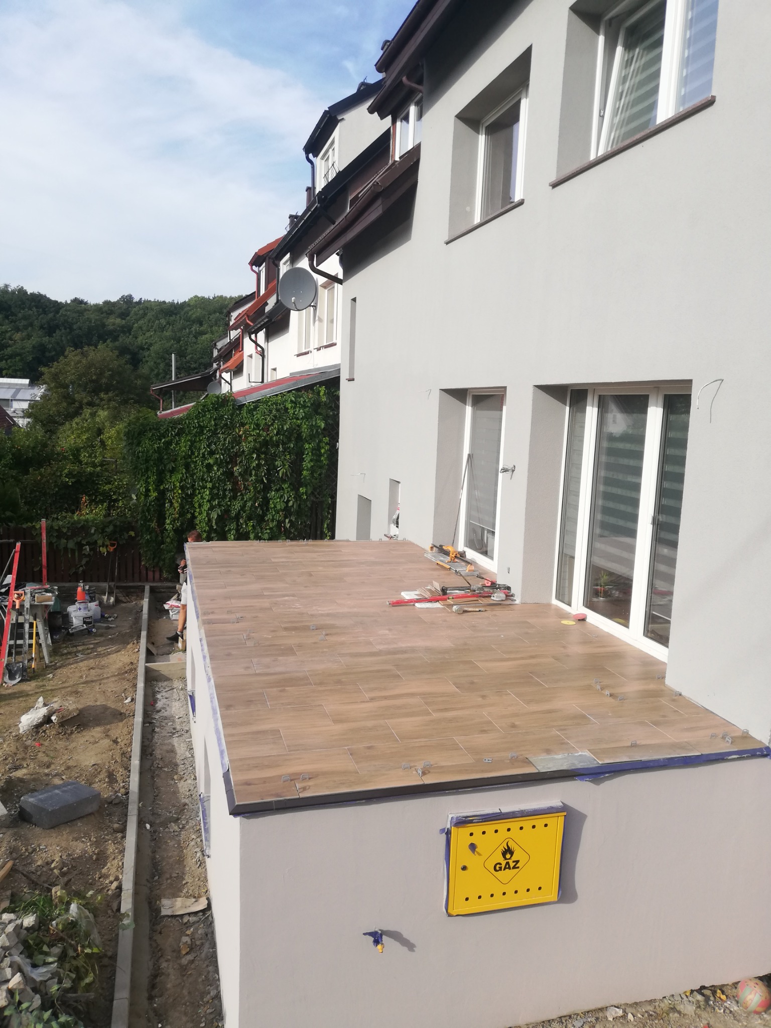 Tarasy zewnętrzne w trakcie wykańczania, płytki imitujące drewno ułożone na wylewce, widoczna skrzynka gazowa na ścianie budynku.