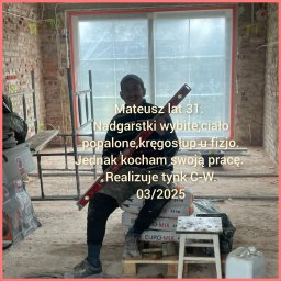 TynkNaturalnie - Uśmiechnięty pracownik budowlany siedzi na workach z zaprawą, trzymając poziomicę na tle surowego wnętrza w trakcie remontu, z widocznymi cegłami i instalacjami.