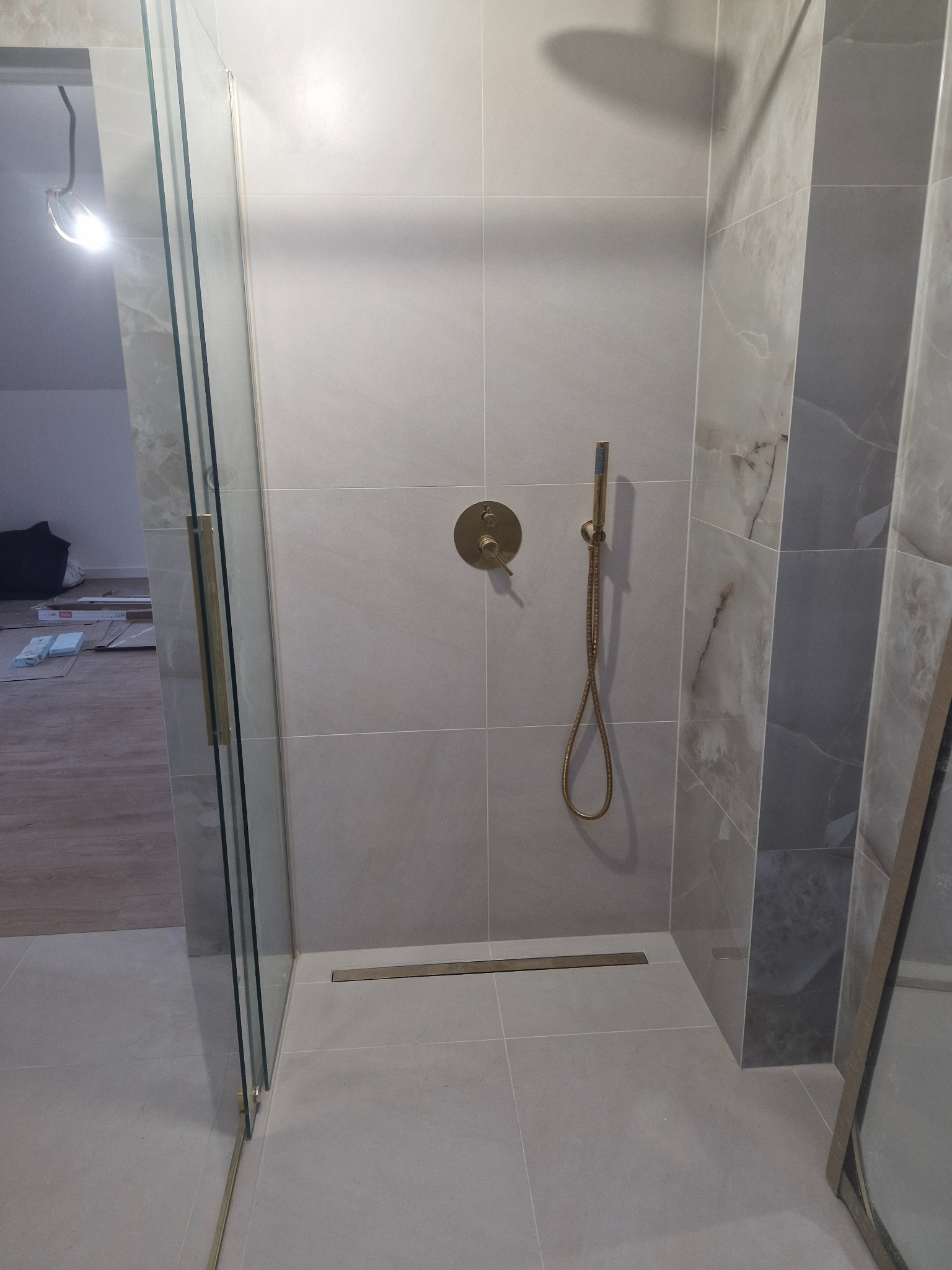 Nowoczesna kabina prysznicowa z szarymi płytkami imitującymi marmur, złotą armaturą i odpływem liniowym; widoczny fragment szklanych drzwi i pomieszczenia w trakcie remontu.