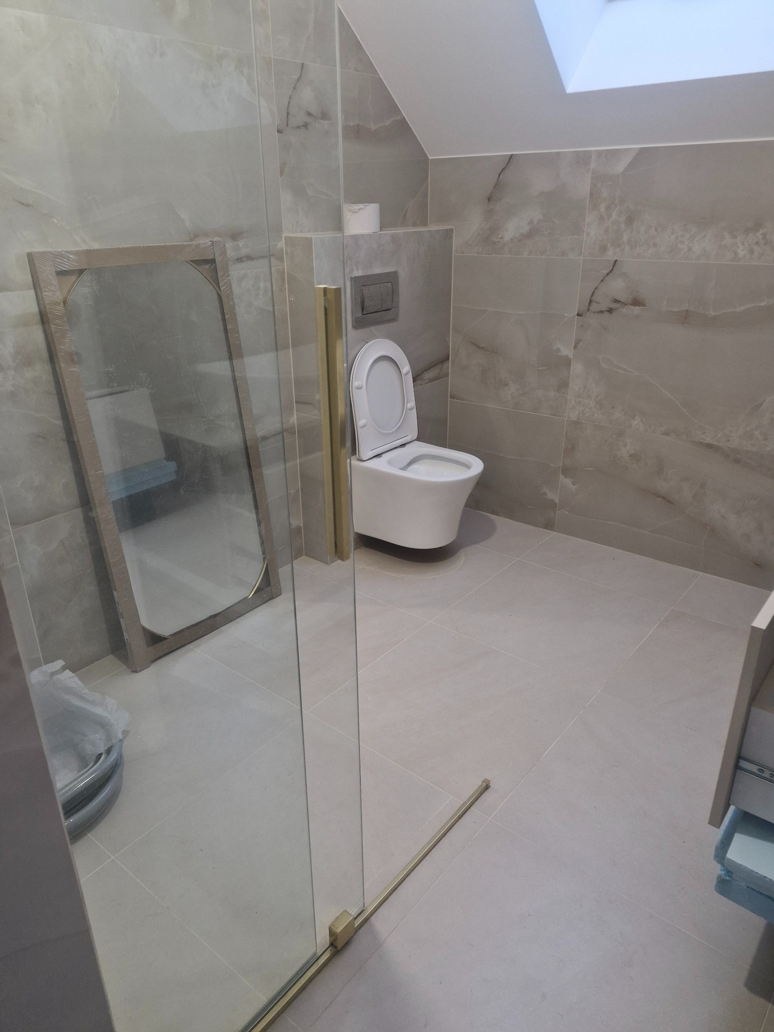 Wykończona łazienka z beżowymi płytkami imitującymi marmur, wisząca toaleta i szklana kabina prysznicowa z złotymi okuciami; lustro oparte o ścianę w folii ochronnej.