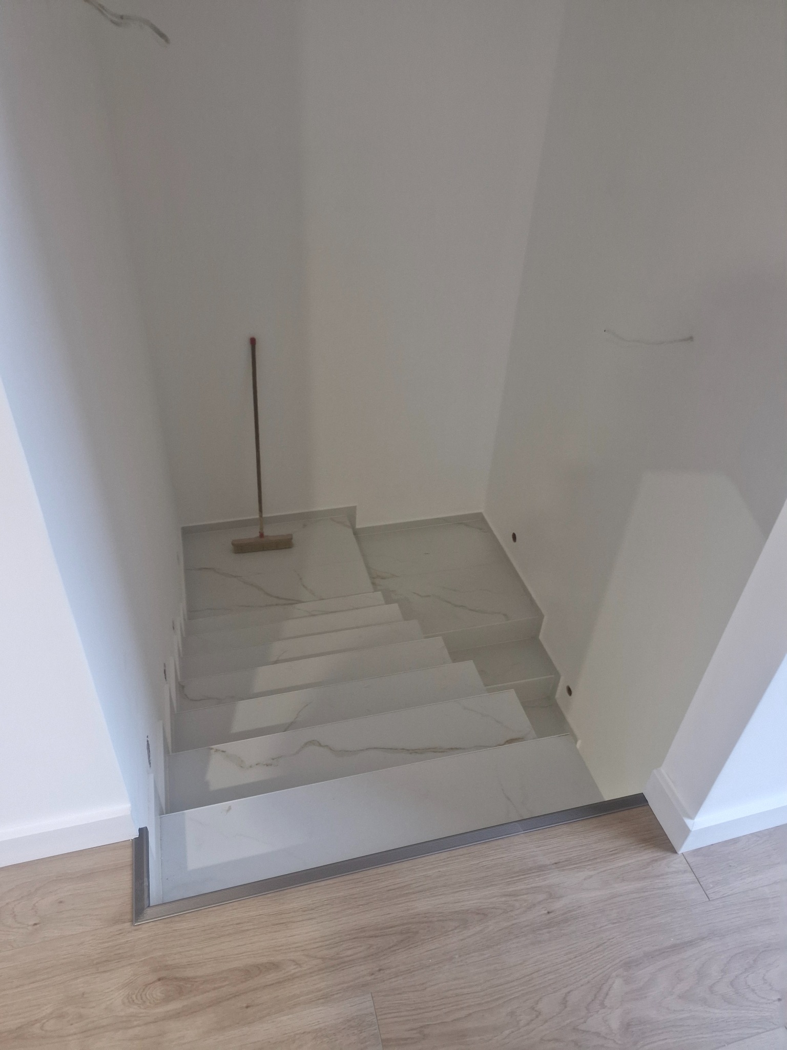 Wykończone schody wewnętrzne z białych płytek imitujących marmur, z widoczną listwą wykończeniową i pozostawionym mopem na podłodze.