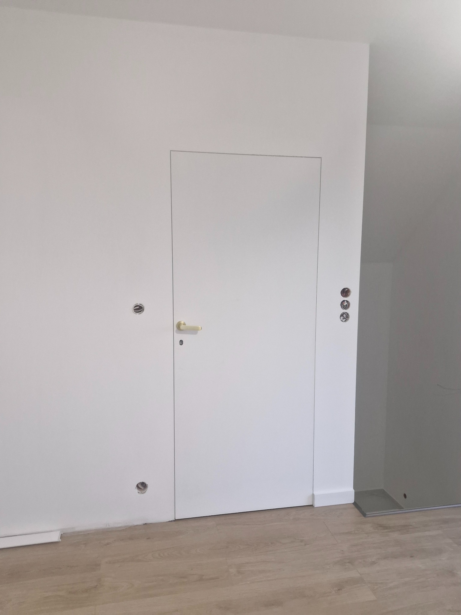 Minimalistyczne wnętrze z ukrytymi drzwiami w białej ścianie, widoczne otwory na gniazdka elektryczne i jasne panele podłogowe.