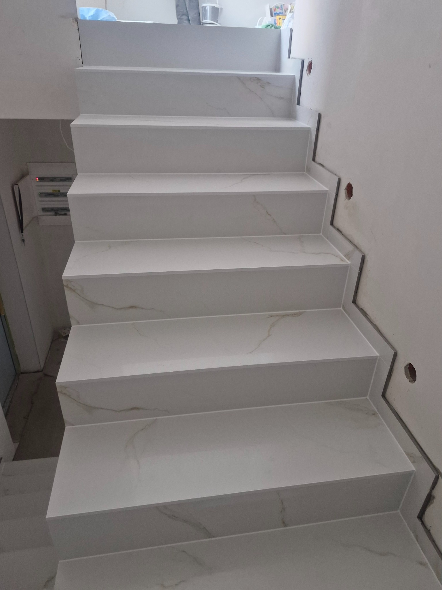 Schody wewnętrzne wykończone białymi płytkami imitującymi marmur, widoczne niedokończone prace wykończeniowe przy ścianie.