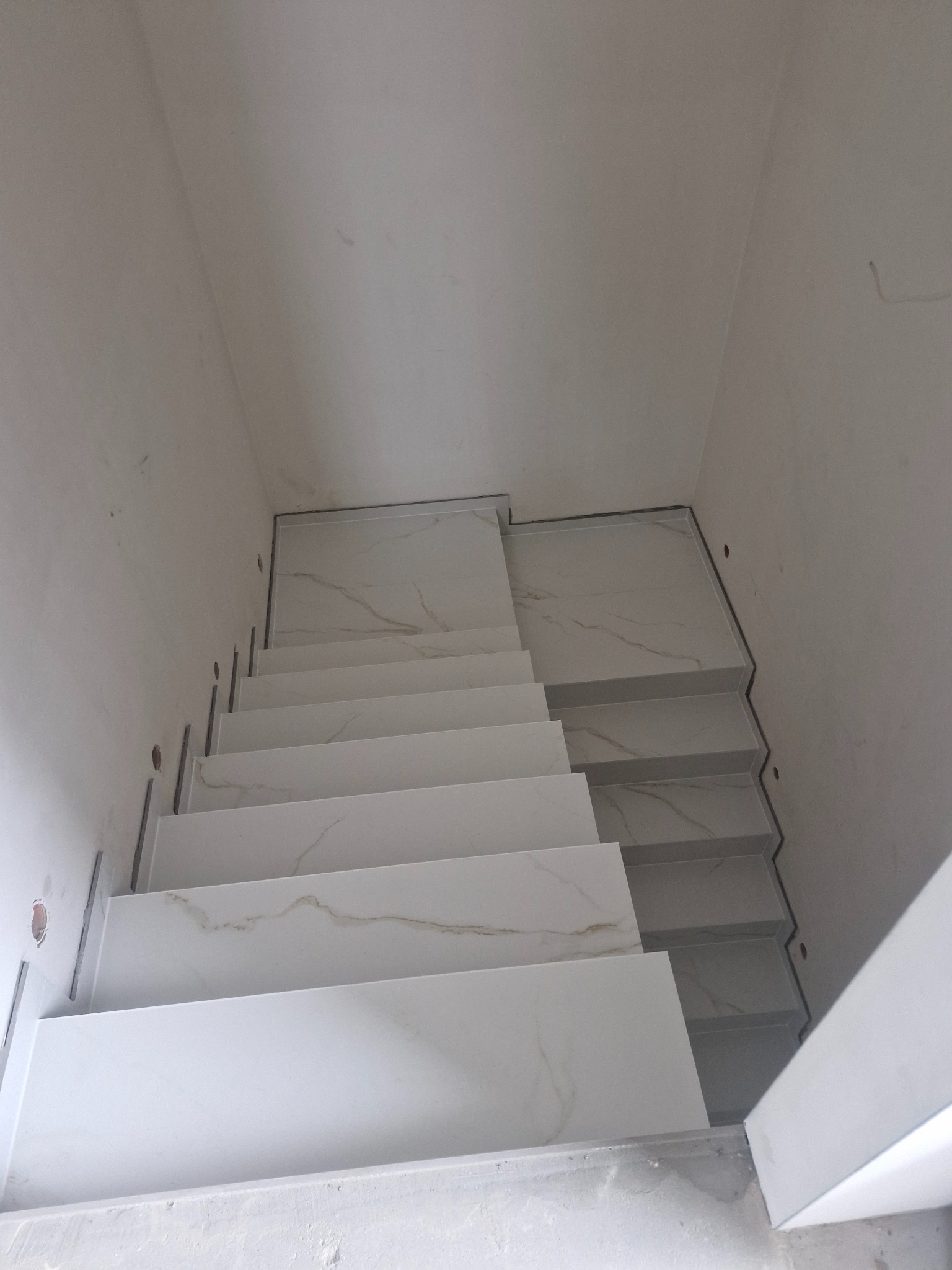 Schody wewnętrzne wyłożone białymi płytkami imitującymi marmur, widoczne otwory montażowe w ścianie, surowy stan wykończenia.