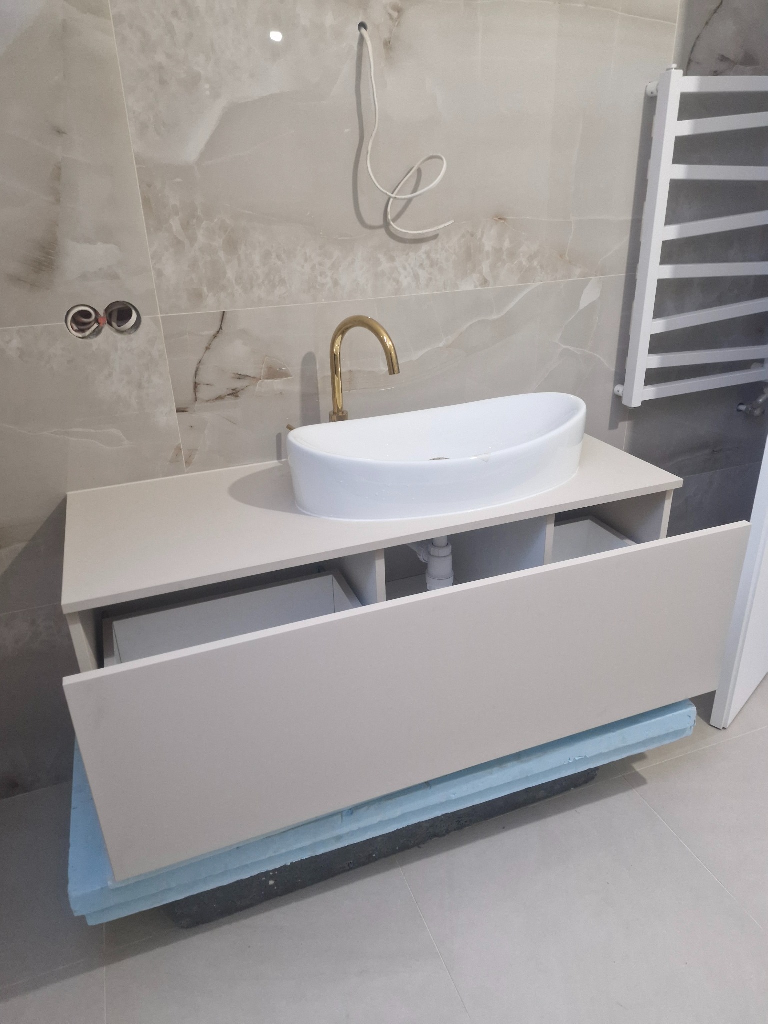 Nowoczesna łazienka w trakcie wykończenia: biała umywalka nablatowa z złotą armaturą, szafka z otwartą szufladą, płytki imitujące marmur na ścianie, grzejnik drabinkowy, widoczne przewody...