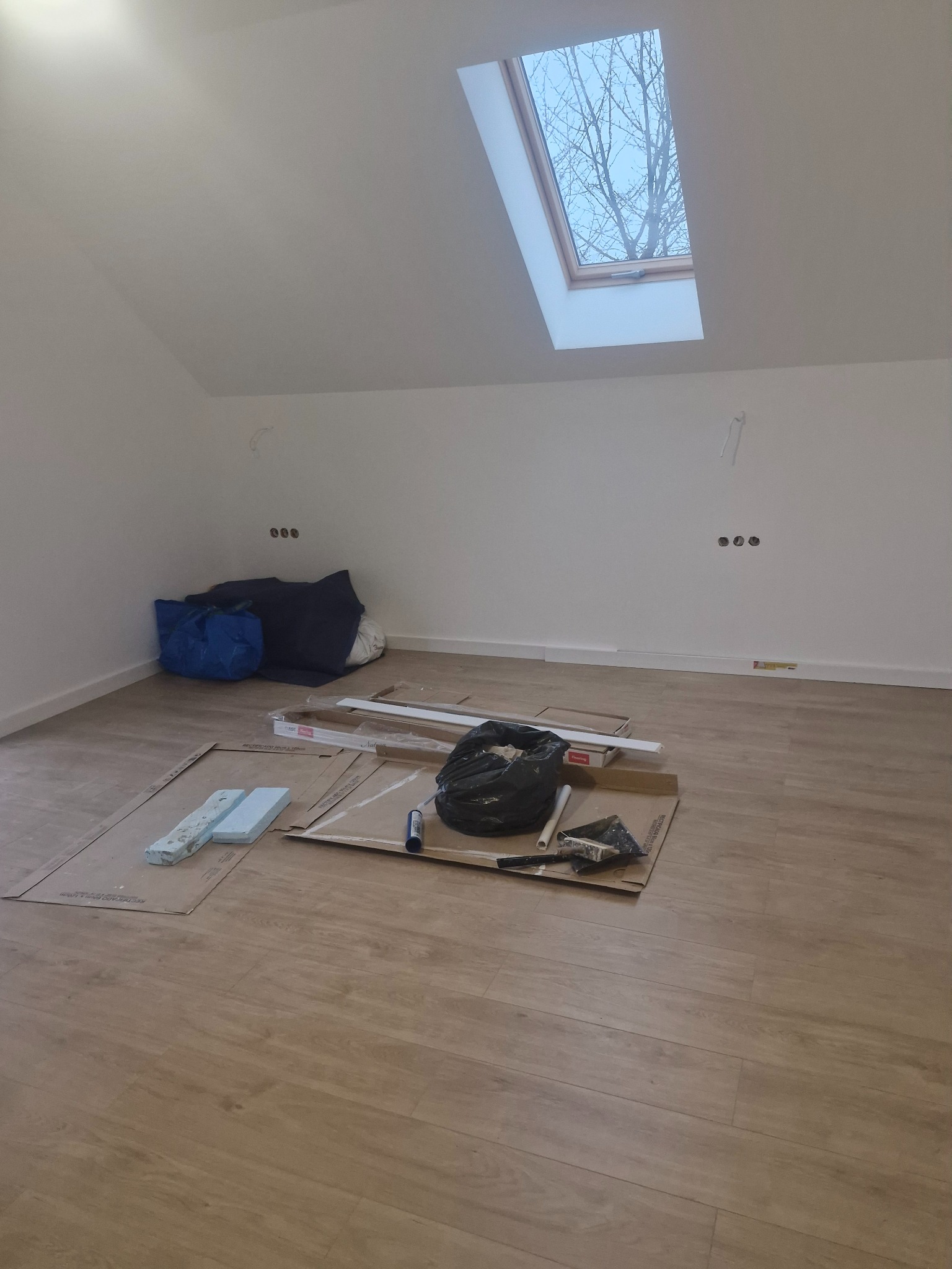 Poddasze w trakcie remontu: widoczne panele podłogowe, narzędzia, worki z materiałami budowlanymi, okno dachowe z widokiem na drzewo bez liści.