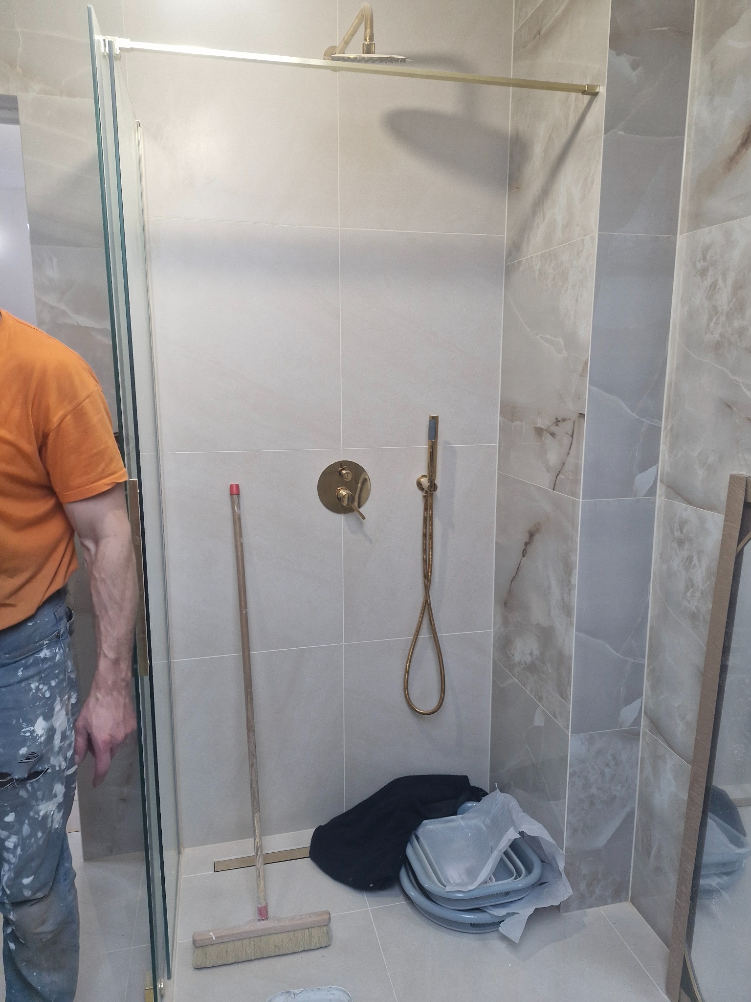 Montaż kabiny prysznicowej z złotą armaturą i szarymi płytkami imitującymi marmur, widoczne narzędzia i ubrudzony farbą pracownik w pomarańczowej koszulce.
