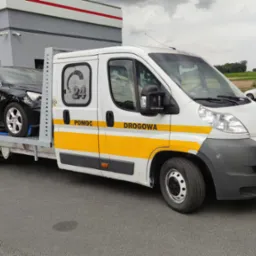 Biała laweta z napisem 'POMOC DROGOWA' i logo 'SERVICE 24' przewozi uszkodzony czarny samochód osobowy na tle stacji benzynowej SPAR express.