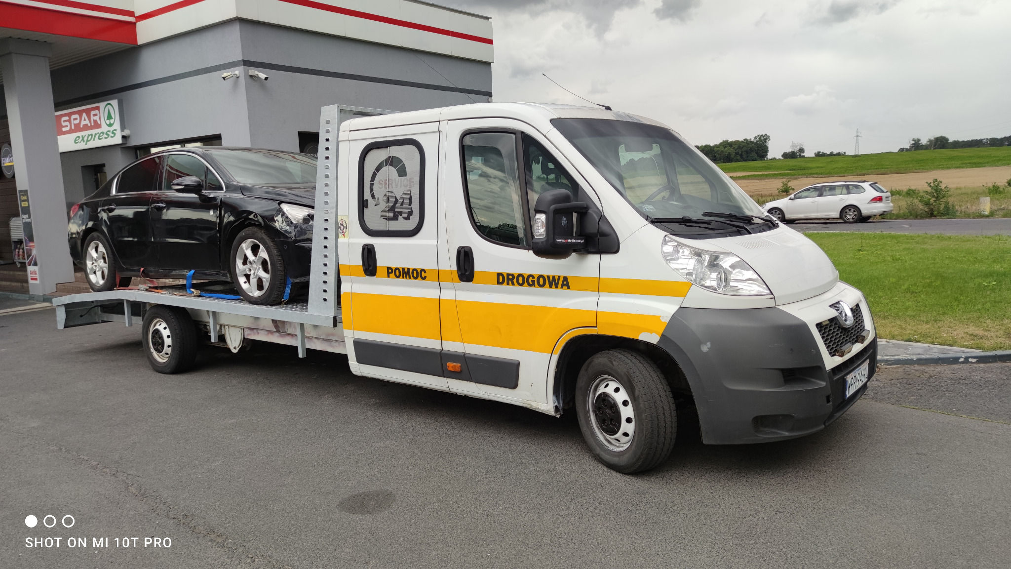 Biała laweta z napisem 'POMOC DROGOWA' i logo 'SERVICE 24' przewozi uszkodzony czarny samochód osobowy na tle stacji benzynowej SPAR express.