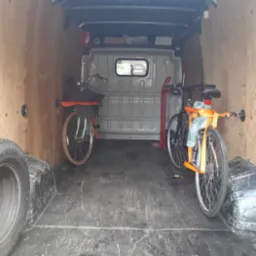 Wnętrze furgonetki z dwoma rowerami zabezpieczonymi pomarańczowymi pasami transportowymi, dodatkowo opony i wózek transportowy.