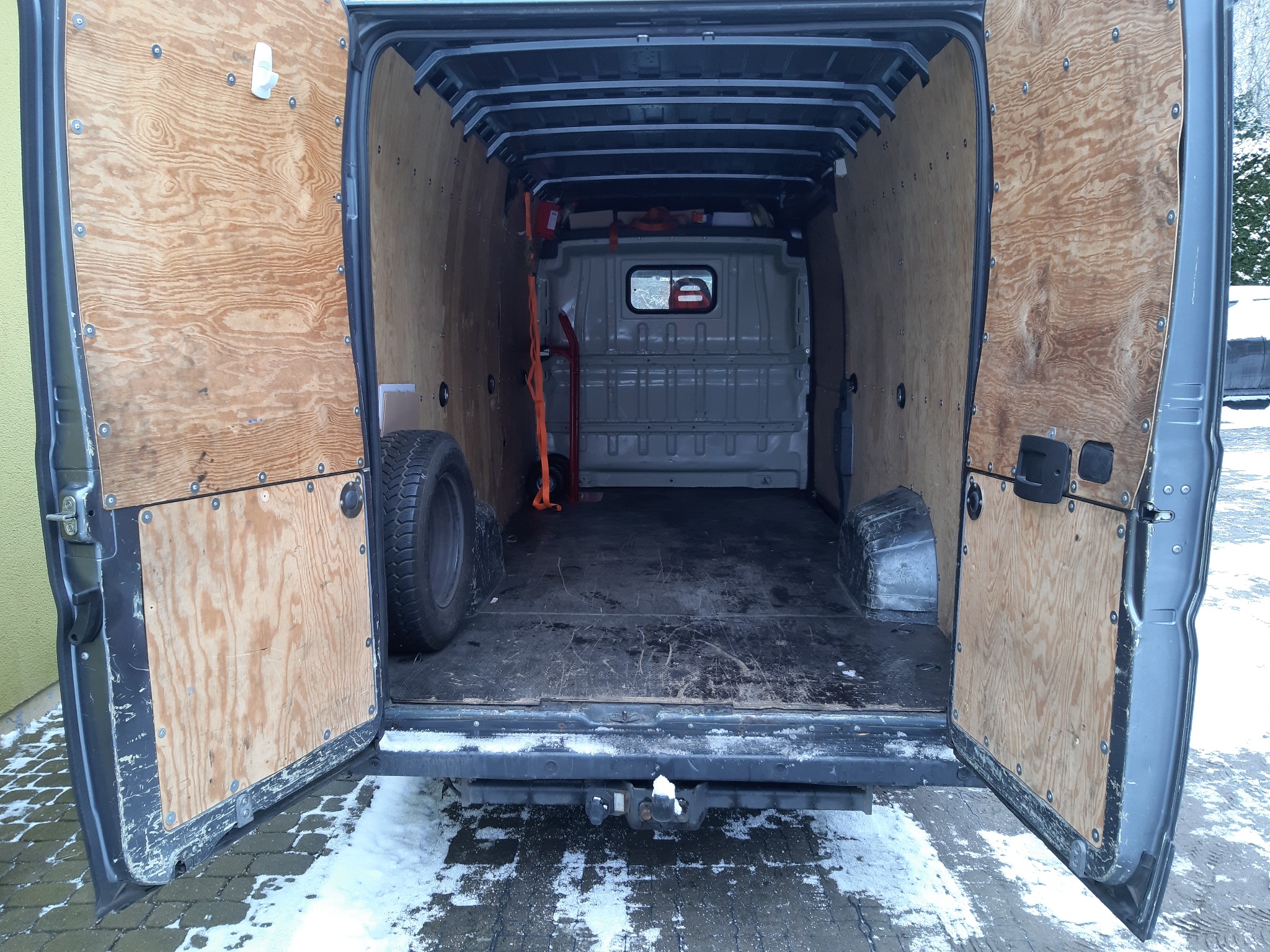 Wnętrze otwartego busa wyłożonego sklejką, z oponą zapasową po lewej stronie i pasami transportowymi wewnątrz, widok z zewnątrz na zaśnieżony podjazd.