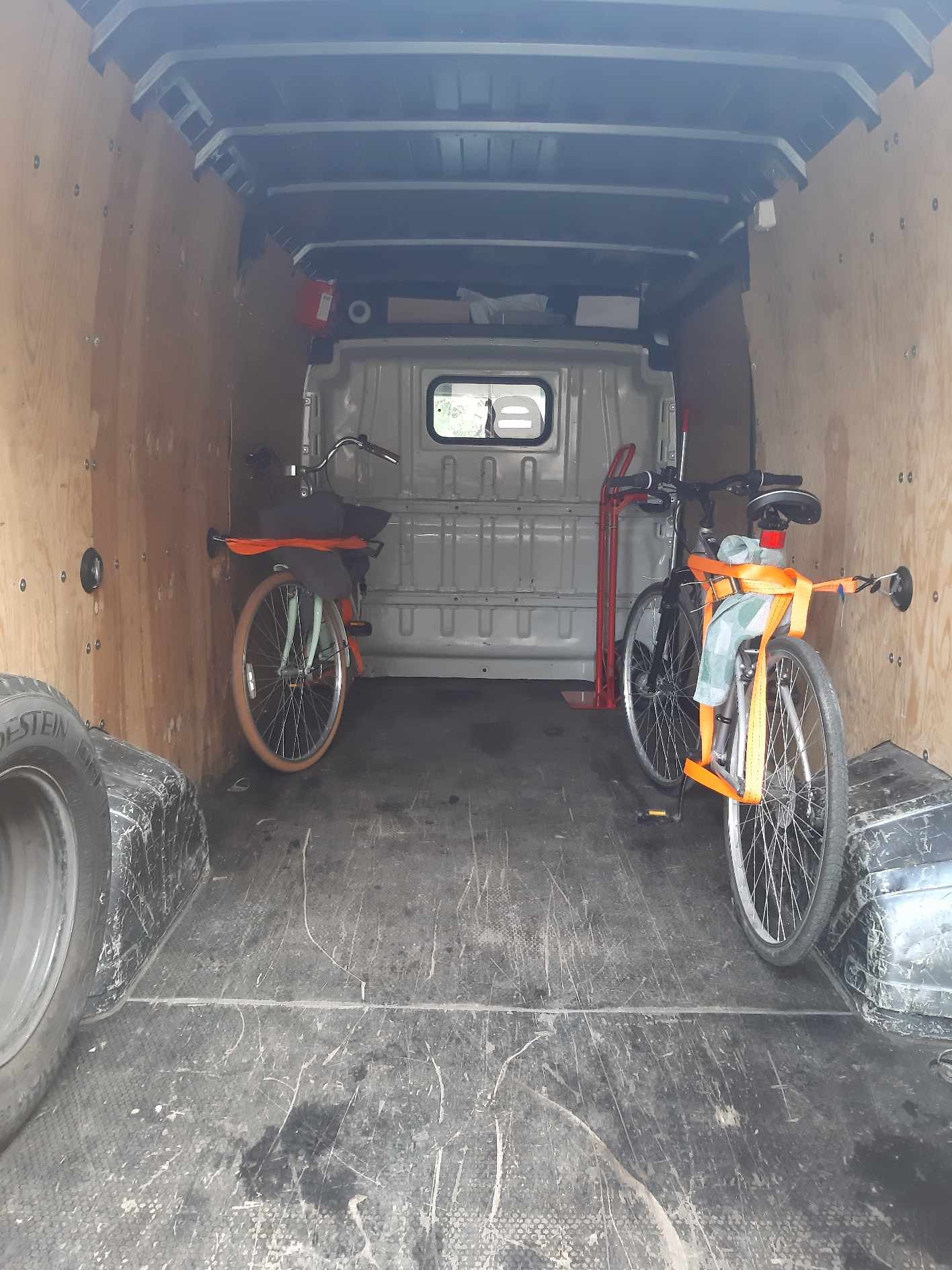 Wnętrze furgonetki z dwoma rowerami zabezpieczonymi pomarańczowymi pasami transportowymi, dodatkowo opony i wózek transportowy.