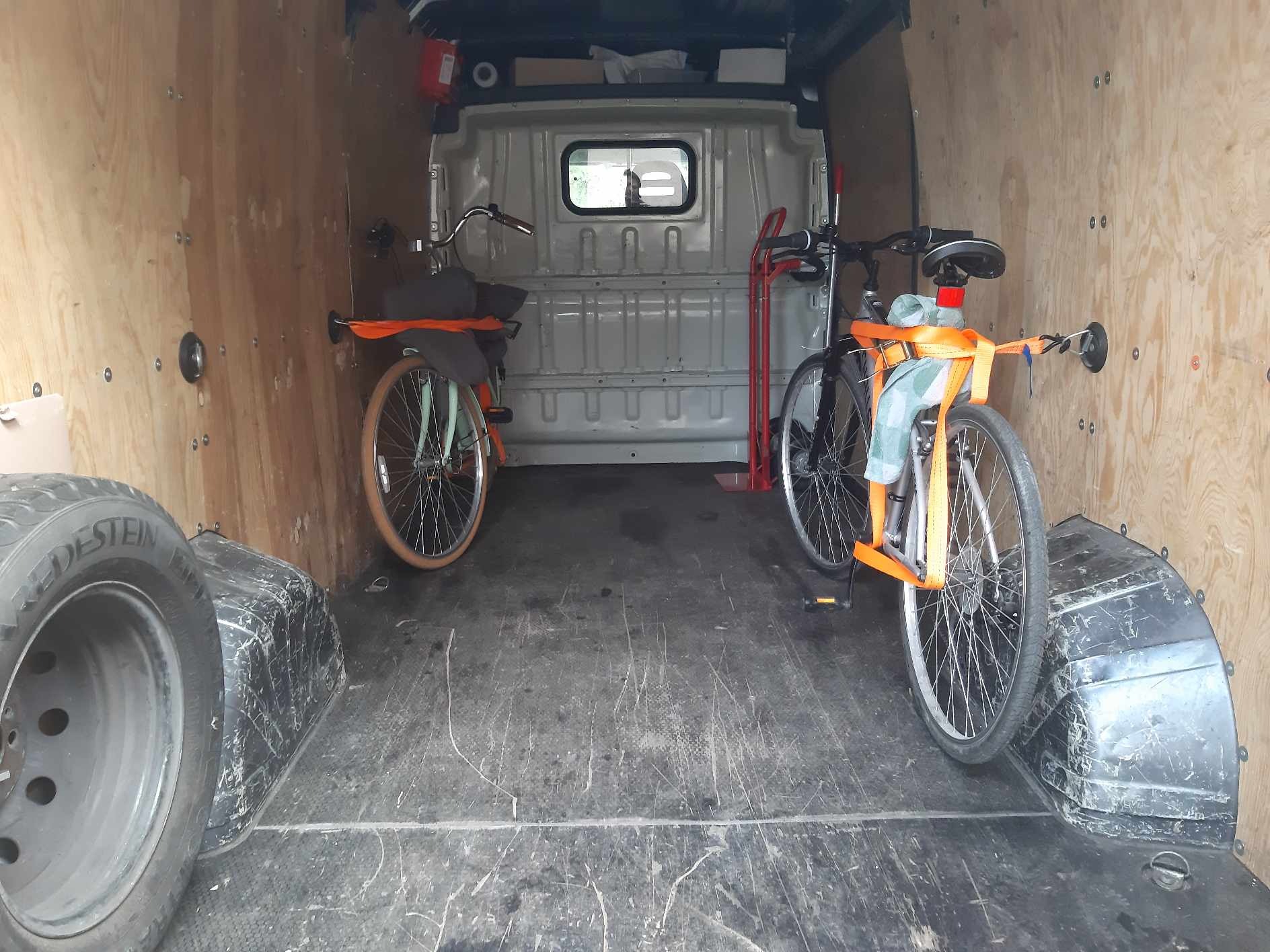 Wnętrze furgonetki z dwoma rowerami zabezpieczonymi pomarańczowymi pasami transportowymi, widoczne także opony i wózek transportowy.
