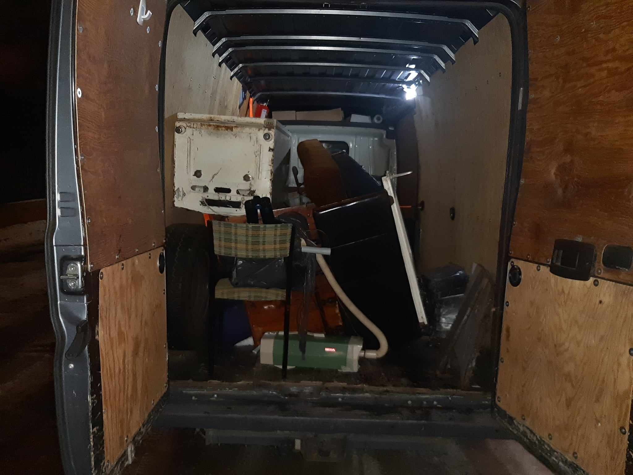 Wnętrze częściowo załadowanego busa z drewnianą zabudową, widoczne różne przedmioty w tym krzesło, kartony i sprzęt czyszczący.