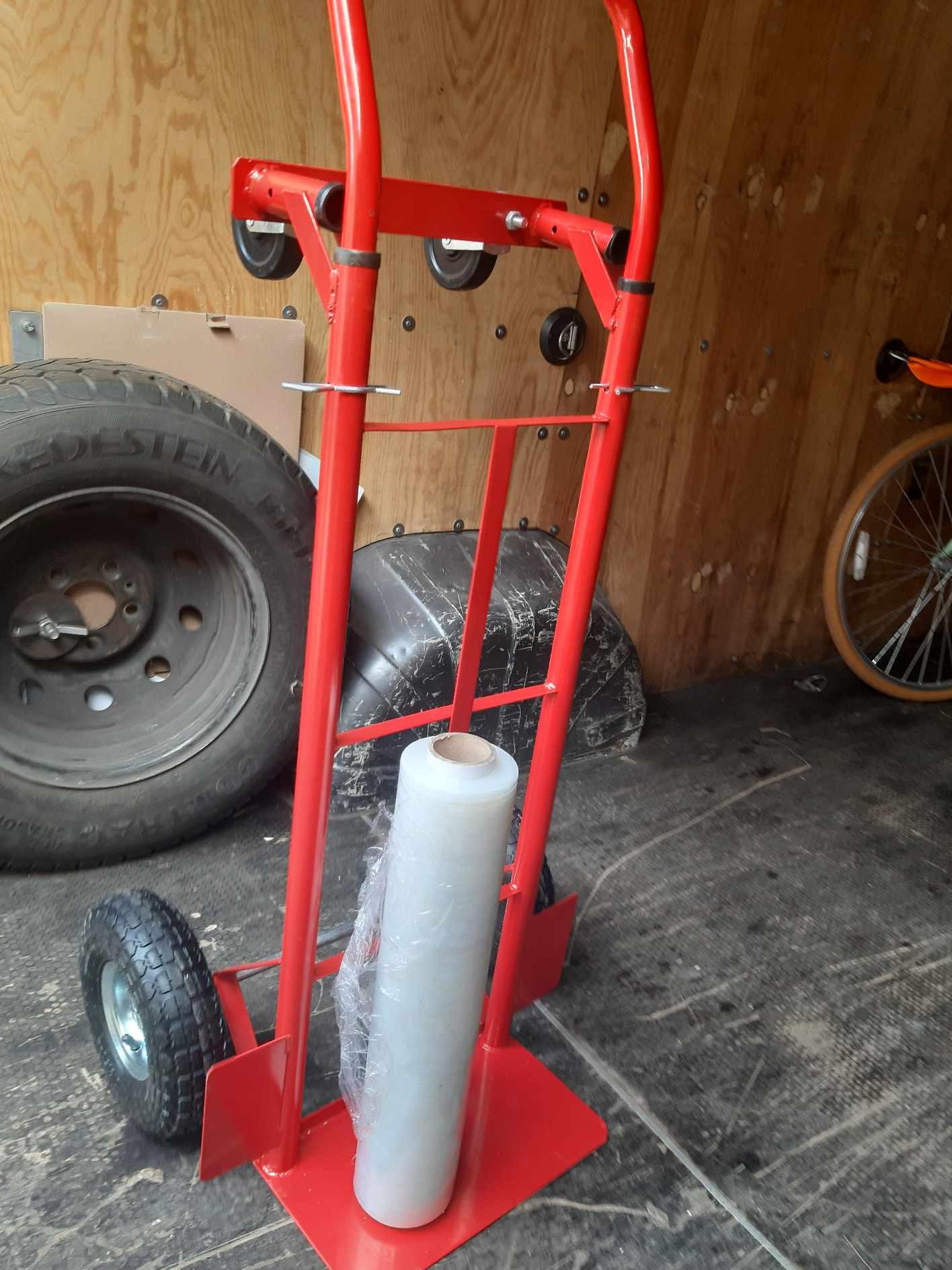 Czerwony wózek transportowy z rolką folii stretch wewnątrz furgonetki, obok opon samochodowych i roweru.
