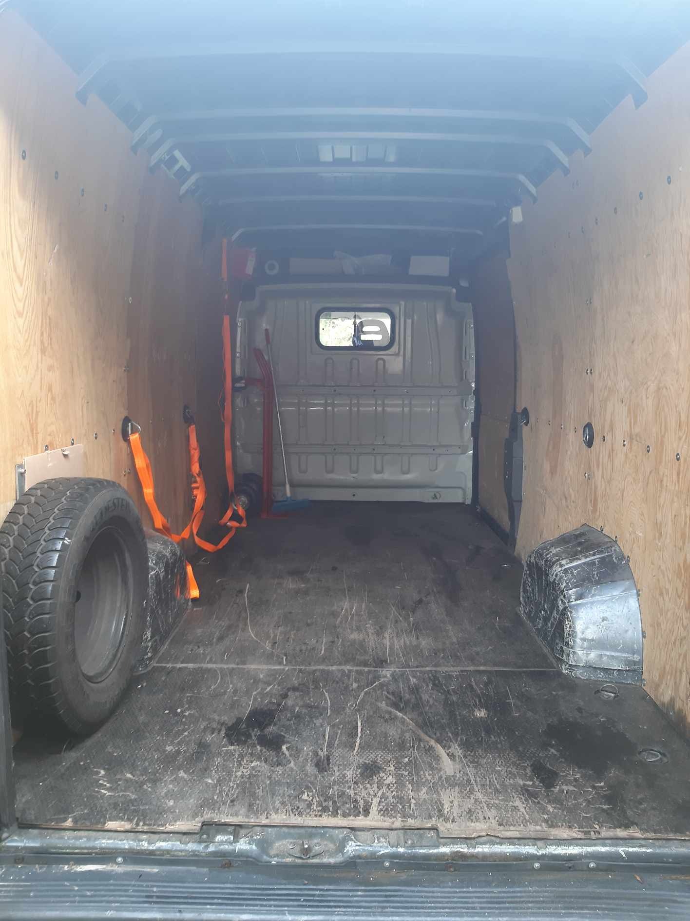 Wnętrze pustej furgonetki z drewnianymi ścianami, widok z tyłu, z zapasowym kołem i pomarańczowymi pasami mocującymi, gotowej do transportu w Aleksandrowie Łódzkim.