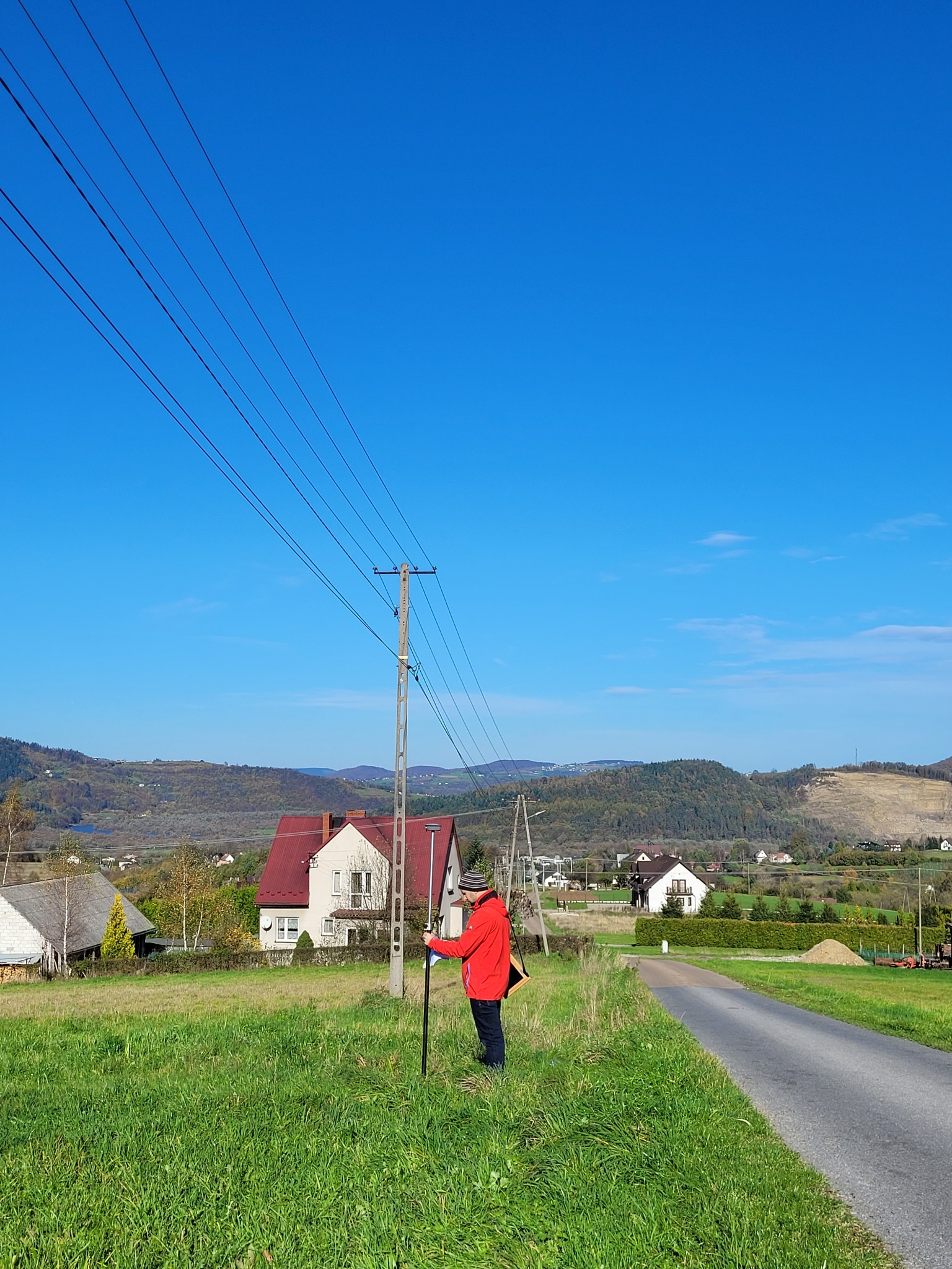 Osoba w czerwonej kurtce i czapce mierzy teren przy drodze za pomocą tyczki geodezyjnej; w tle domy i wzgórza.