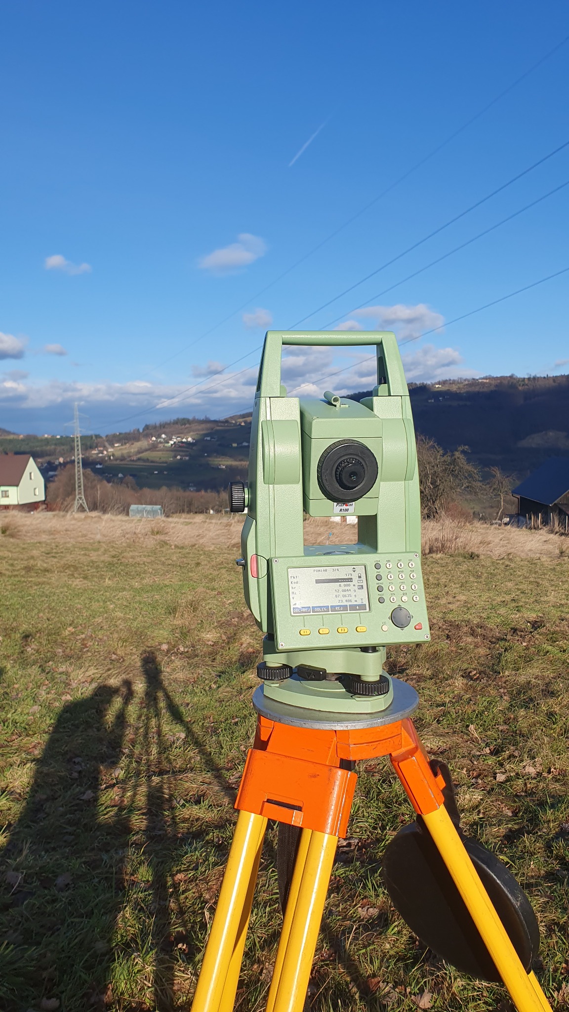 Zielony teodolit elektroniczny na pomarańczowo-żółtym statywie, ustawiony na trawiastym terenie z widokiem na pagórki i błękitne niebo z chmurami i liniami energetycznymi w tle. Widoczny cień statywu.
