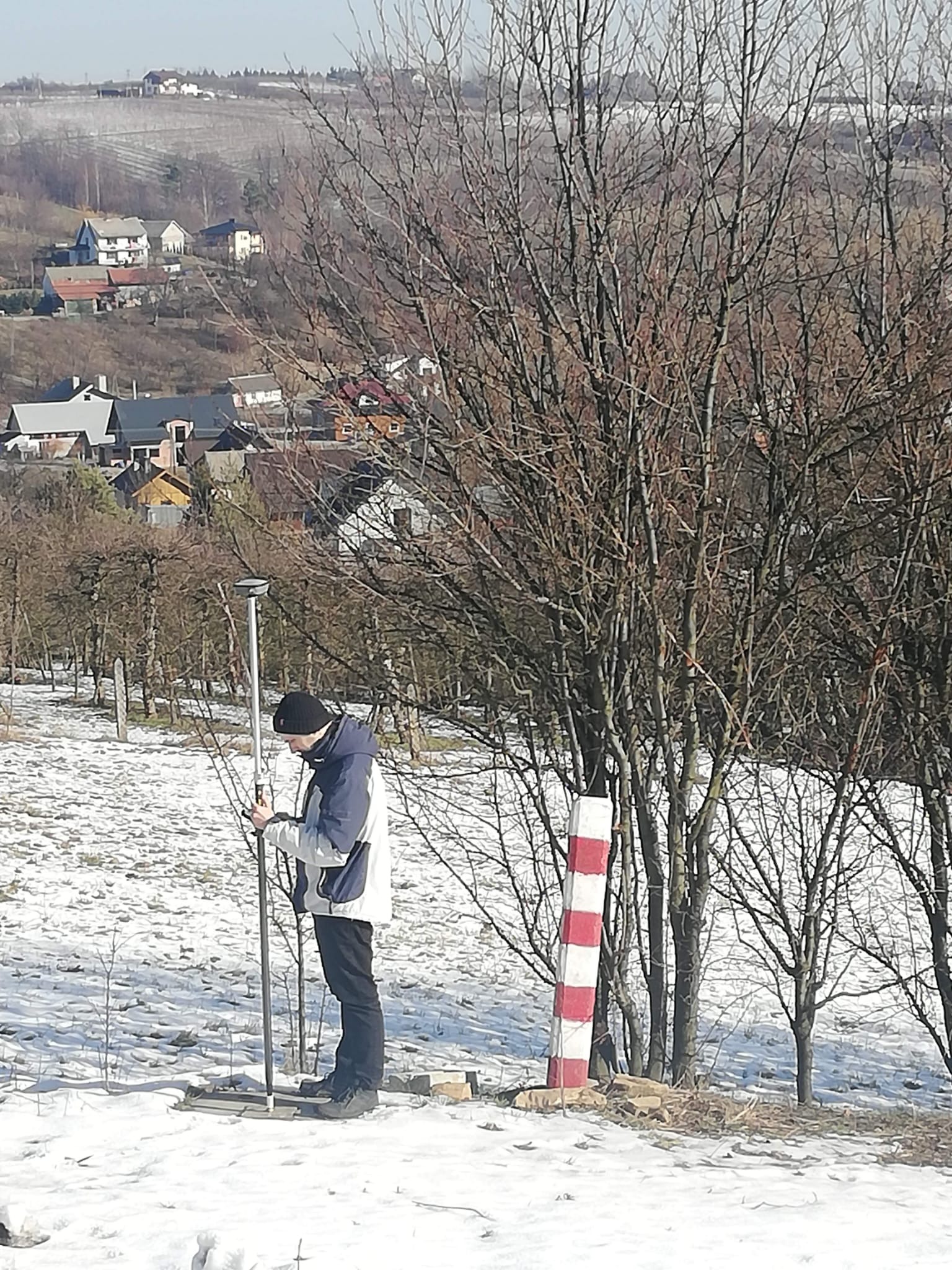 Mężczyzna w zimowej scenerii, ustawia tyczkę geodezyjną na zasnieżonym wzgórzu, w tle zabudowa wiejska i drzewa bez liści.