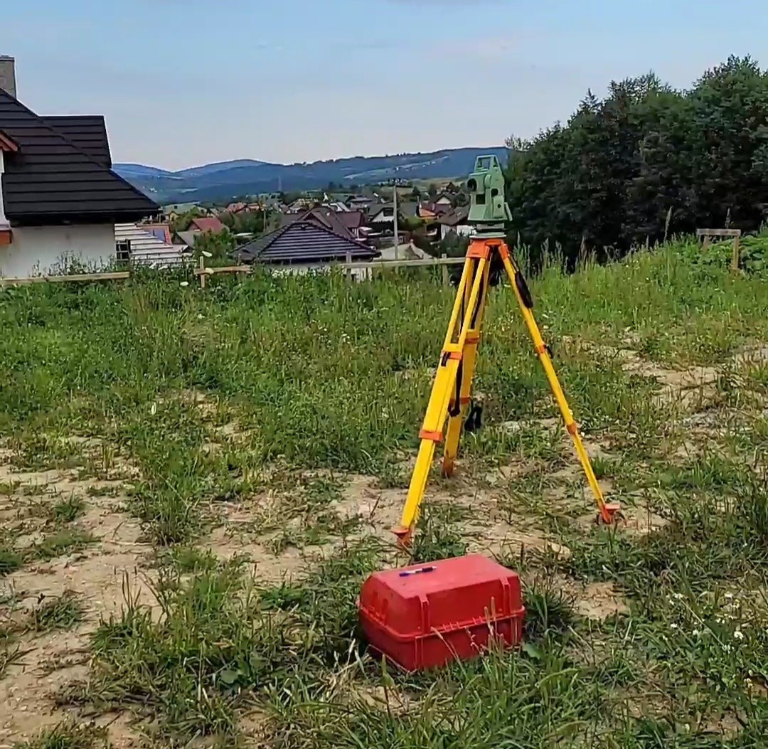 Żółty statyw z teodolitem na zarośniętej działce budowlanej, w tle dom z ciemnym dachem i zabudowania wiejskie.