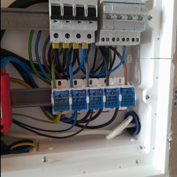 TOMASZ KUŹMA ZETTEC - Szczegółowe ujęcie wnętrza skrzynki elektrycznej z zainstalowanymi bezpiecznikami, przewodami i listwami zaciskowymi. Widoczne elementy marki Eaton i Hager.