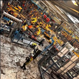 TOMASZ KUŹMA ZETTEC - Rozległa hala fabryczna z licznymi żółtymi robotami przemysłowymi; pracownicy w kamizelkach i kaskach przy stanowiskach. Widok z góry, perspektywa zniekształcona.