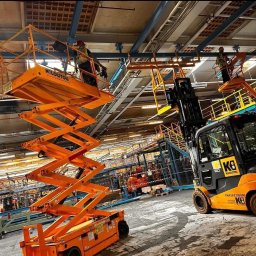 TOMASZ KUŹMA ZETTEC - Praca w hali: elektryk na podnośniku montuje oświetlenie pod sufitem. W tle widoczny wózek widłowy i inne podnośniki. Drobne prace na wysokości.