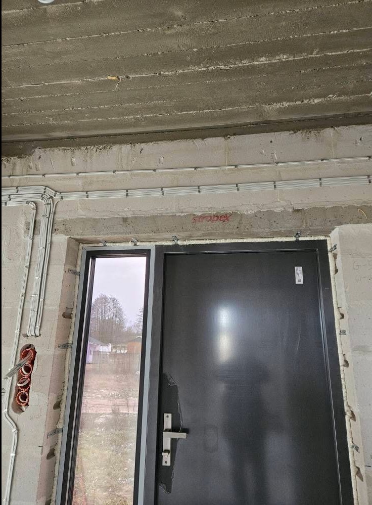 Surowy stan: szara, metalowa ościeżnica drzwiowa z szybą, widoczne przewody elektryczne w białych listwach na ścianie z betonu komórkowego i suficie.