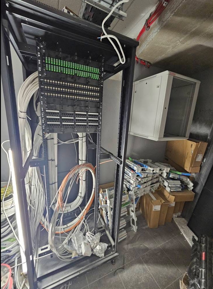 Szafa rack z panelami krosowymi i okablowaniem w trakcie montażu. Widoczne elementy instalacji i skrzynka elektryczna na ścianie.